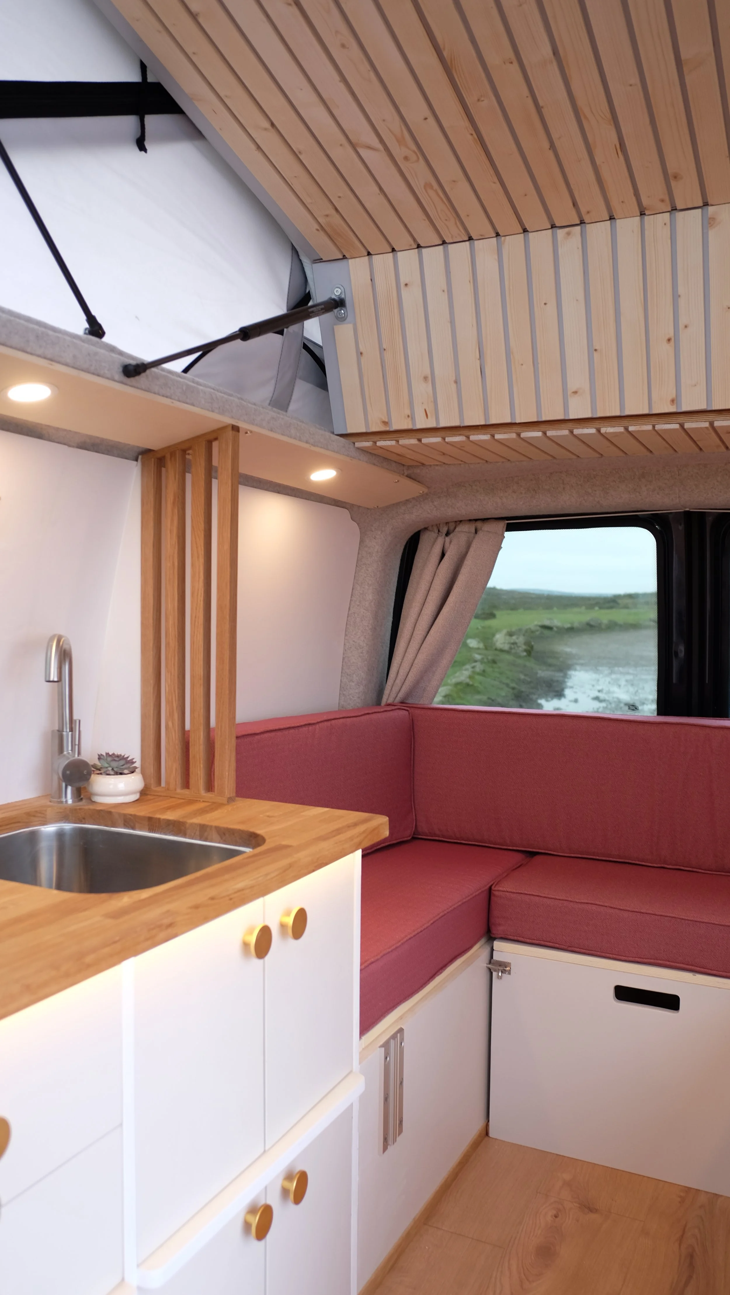 Interior of a camper van with a small kitchen area, red cushioned seating, and wooden accents. A window shows a view of a river and green landscape.