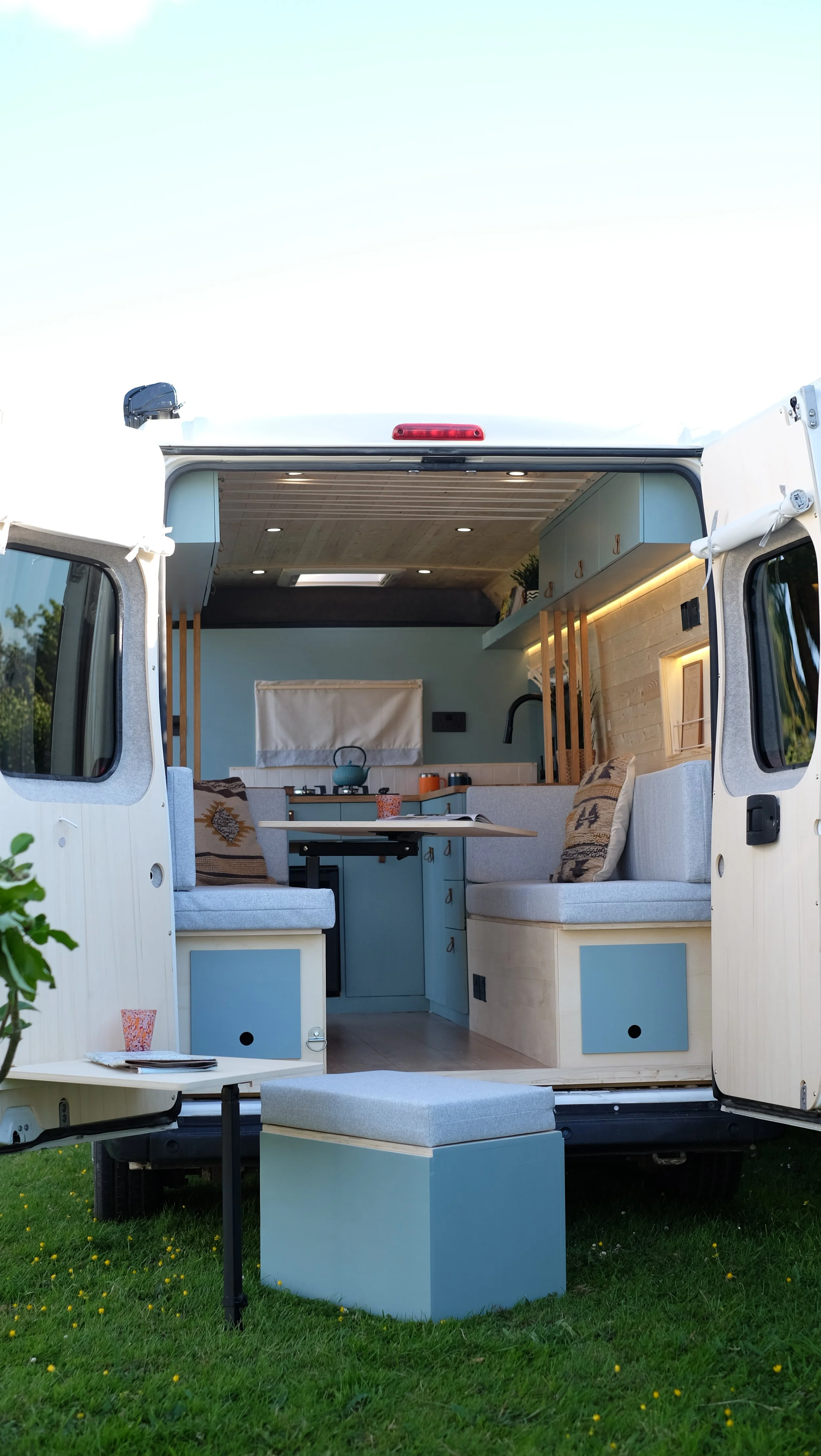 Interior of a customized camper van with a seating area, kitchen, and small table on a grassy area outside.