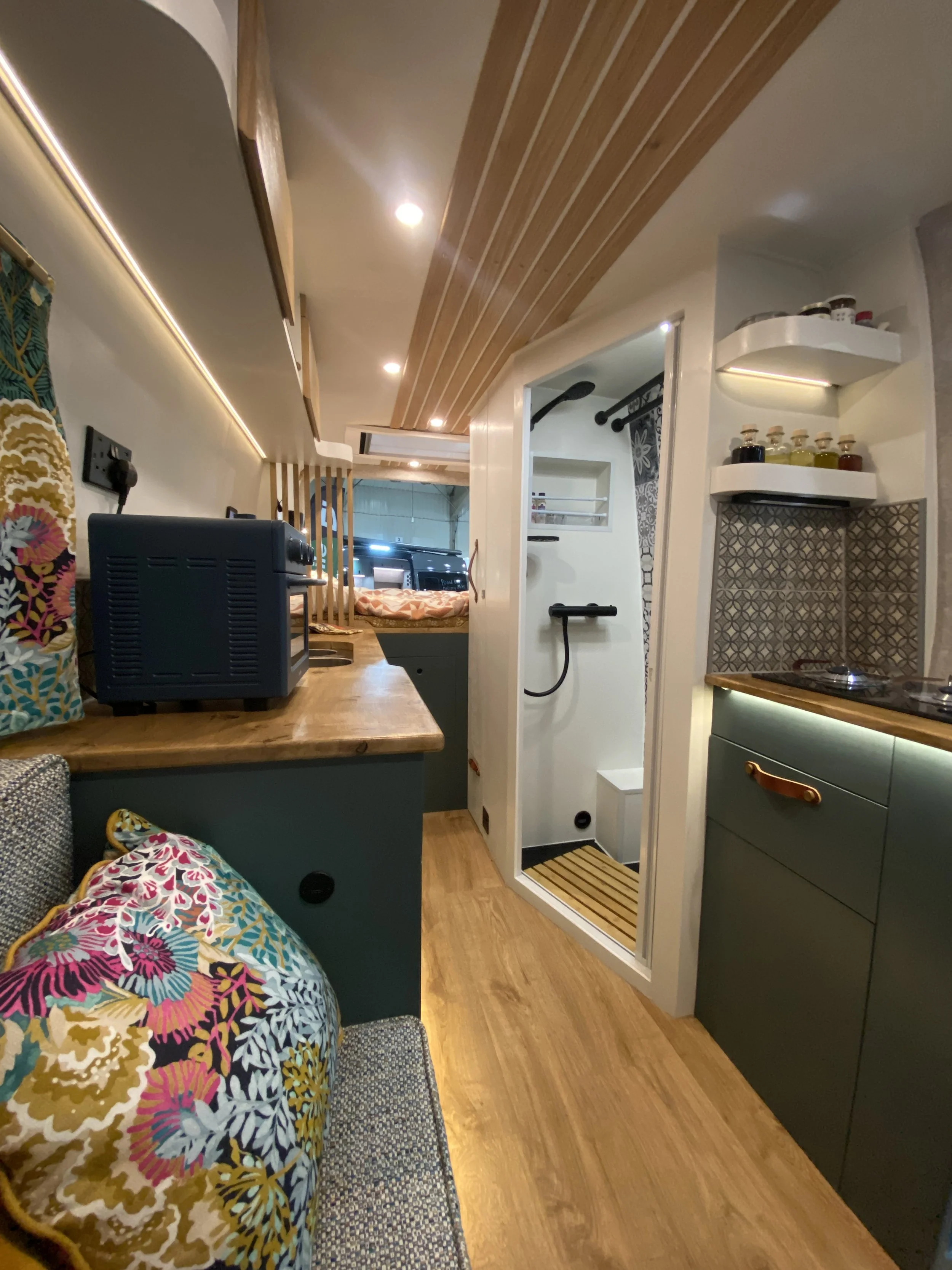 Interior of a small living space with a kitchenette, a colorful patterned pillow, a wood countertop, a shower stall, and a bedroom area in the background.