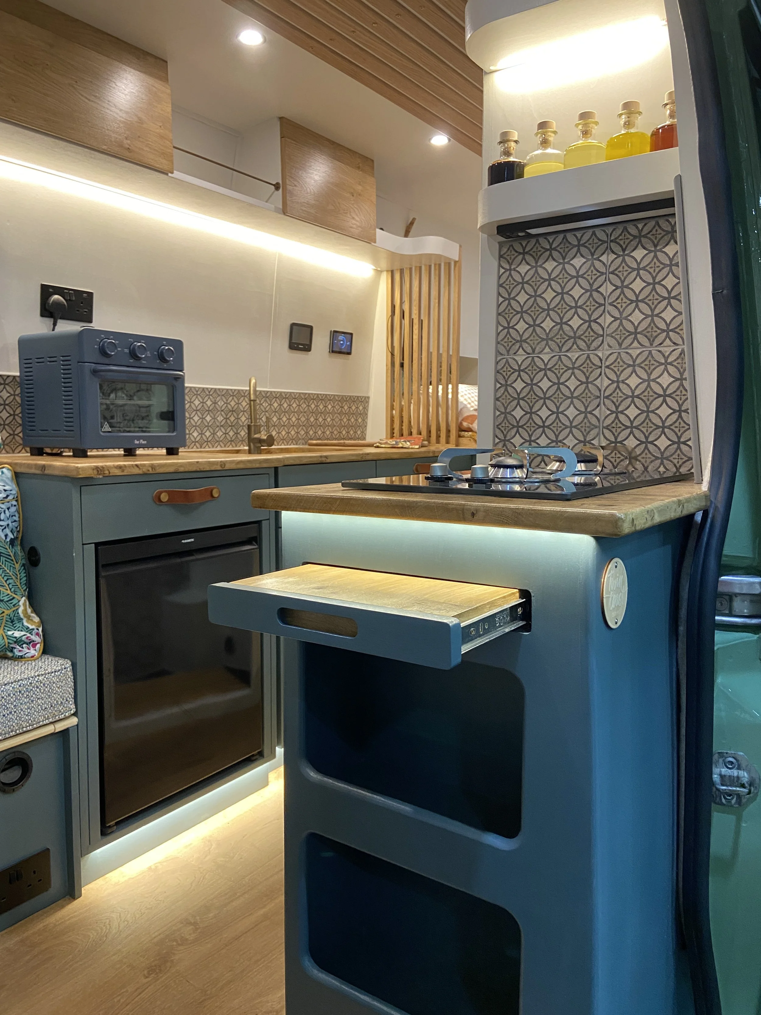 Cozy compact kitchen area with a blue mini oven, stovetop, drawer, and decorative tile backsplash, set in a modern, stylish tiny home or camper with wooden accents and subtle lighting.
