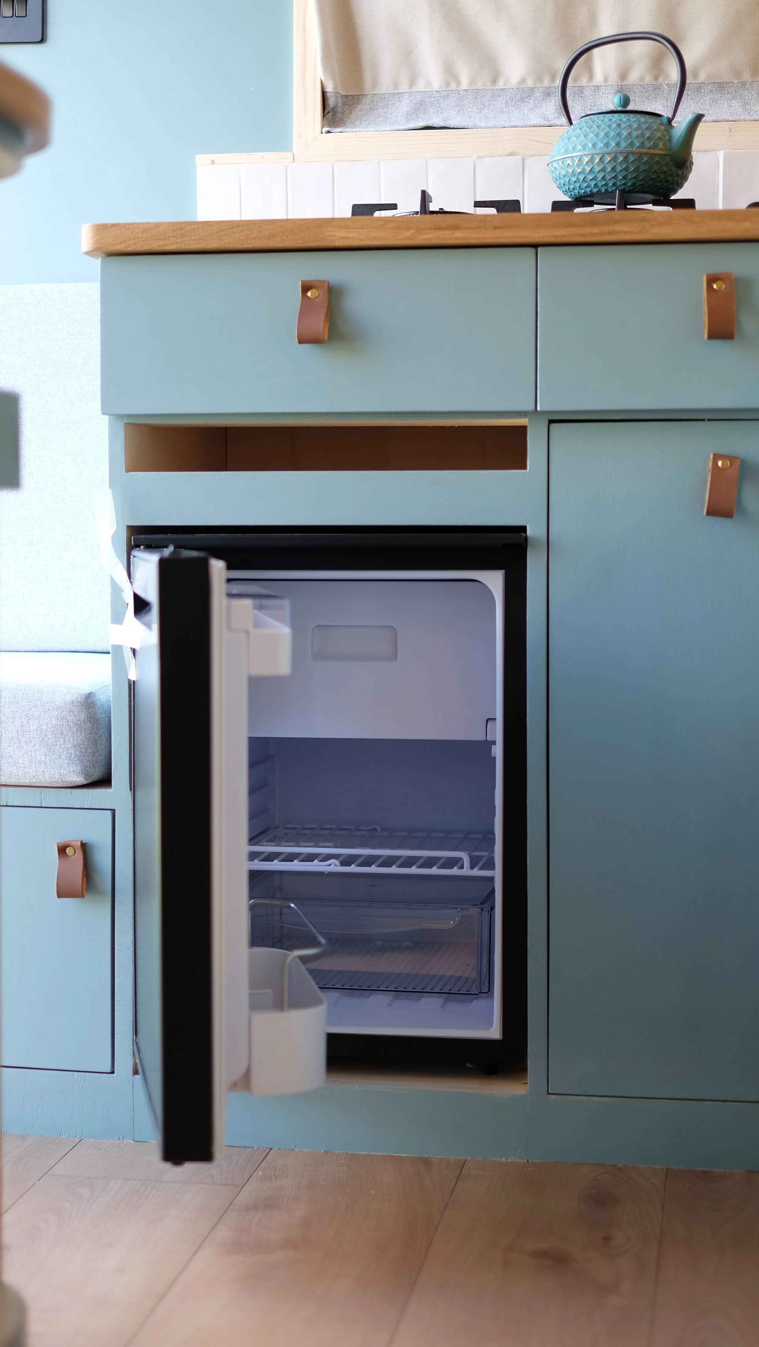 Open small refrigerator with empty interior in a pastel blue kitchen cabinet.