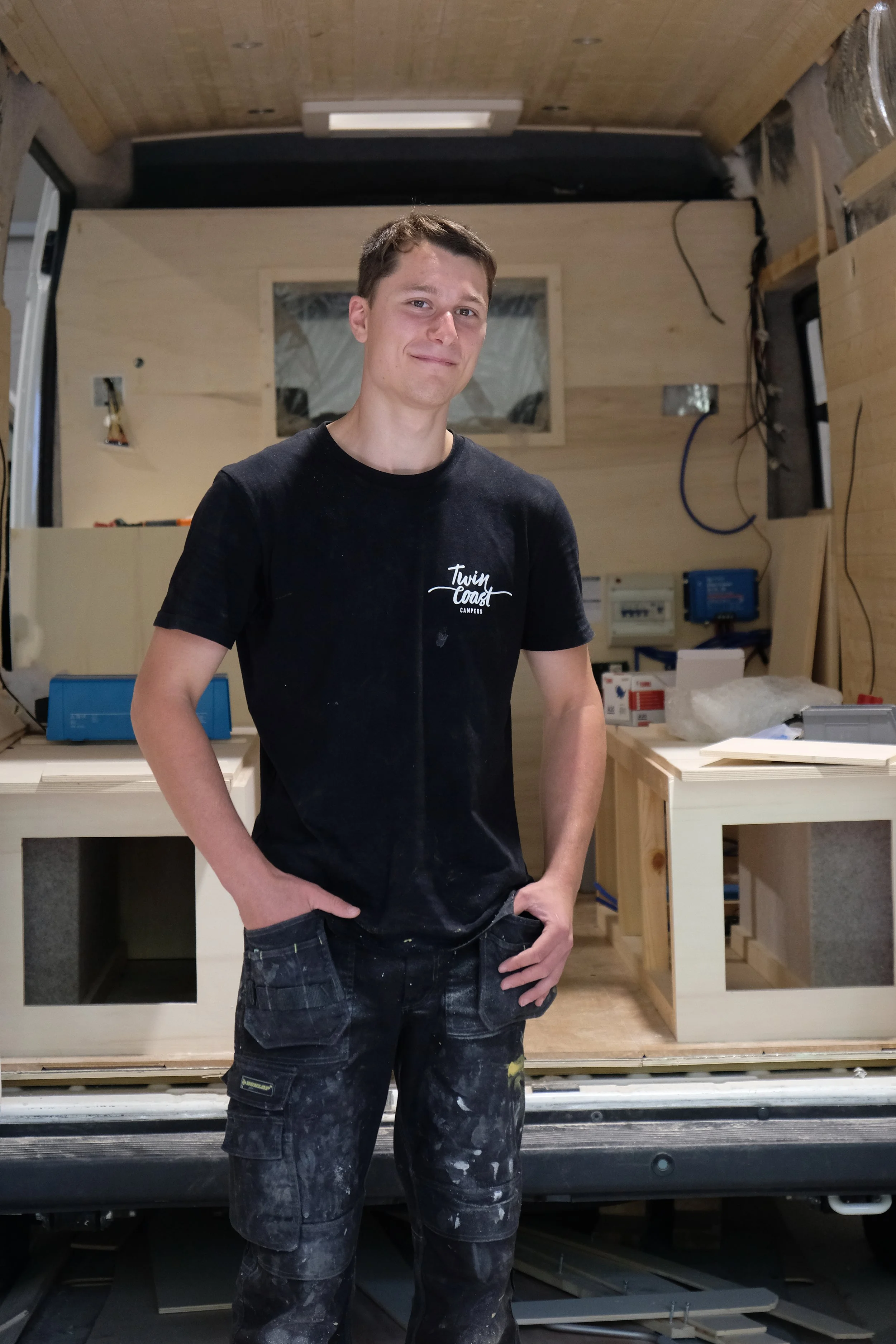 Young man standing inside a camper van under construction, wearing a black T-shirt and paint-splattered work pants, with a partially built wooden interior behind him.