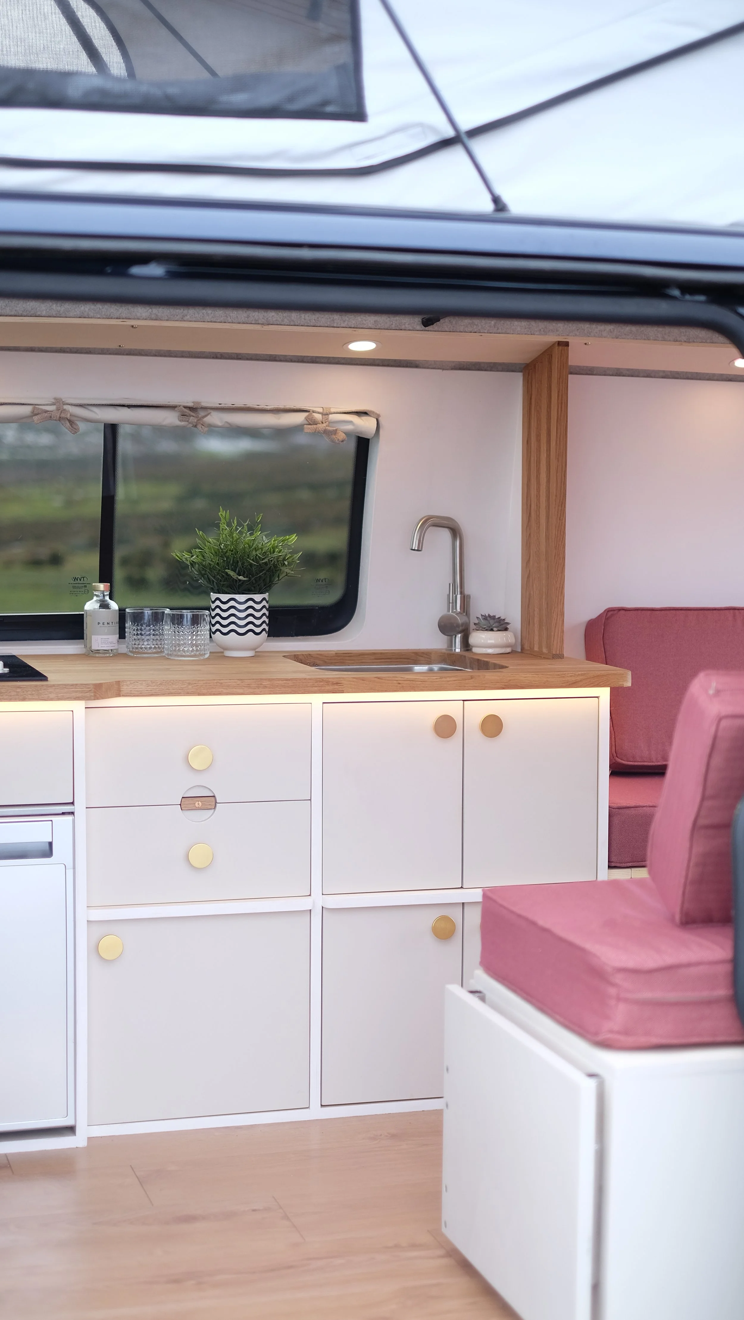 Interior of a compact, modern camper van kitchen with white cabinets, a wooden countertop, a window with a landscape view, a potted plant, drinking glasses, a small bottle, a sink with a faucet, and pink seating cushions.