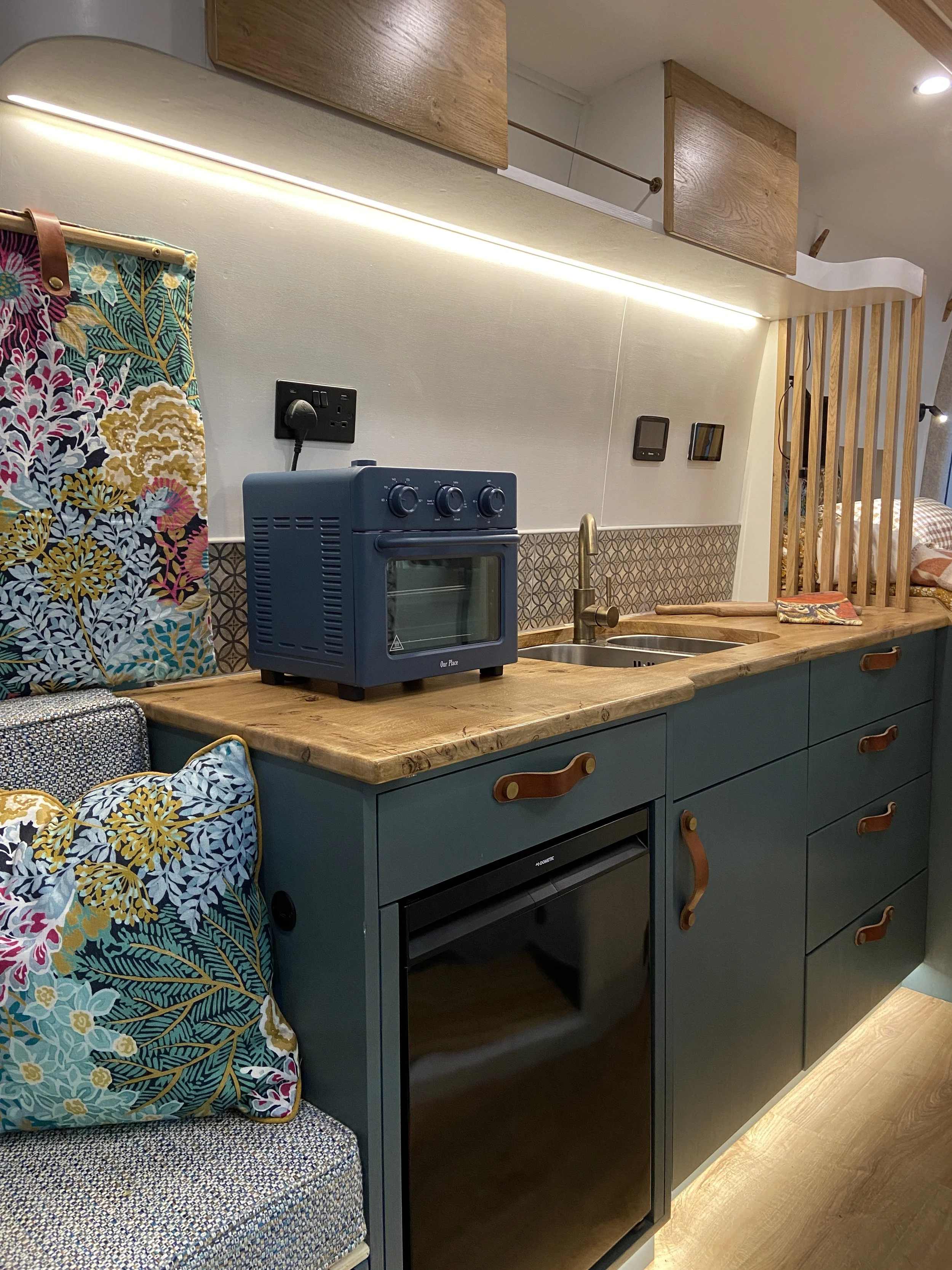 Compact kitchen area with teal cabinetry, a wooden countertop, and a small oven, under warm LED lighting. Decor includes a colorful fabric pillow with a floral pattern on a small floral fabric chair, and a paper bag with a colorful design on the coun