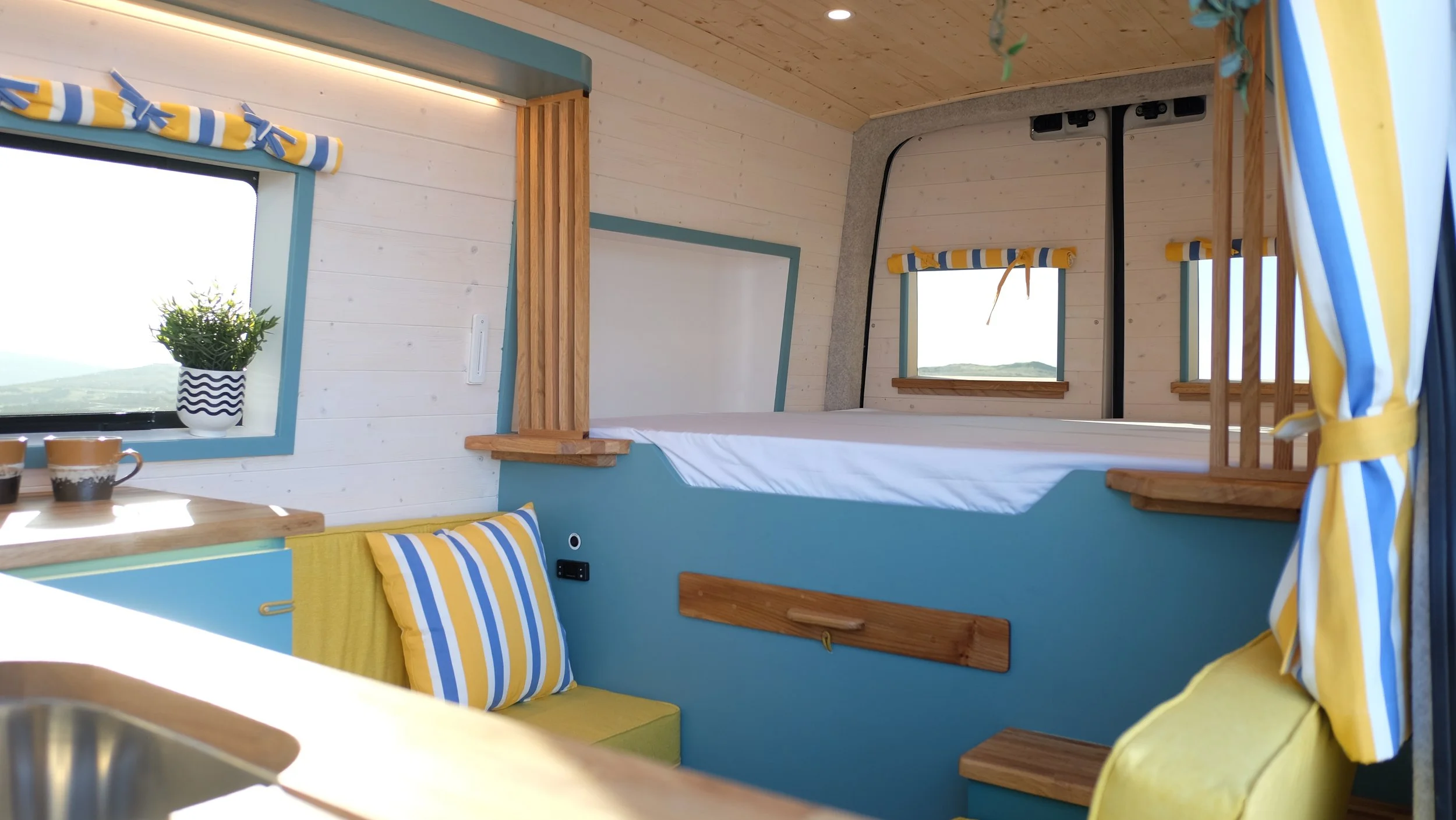 Interior of a small, cozy camper or tiny home with blue and yellow decor, featuring a bed elevated above a seating area, windows with striped curtains, and wooden accents.