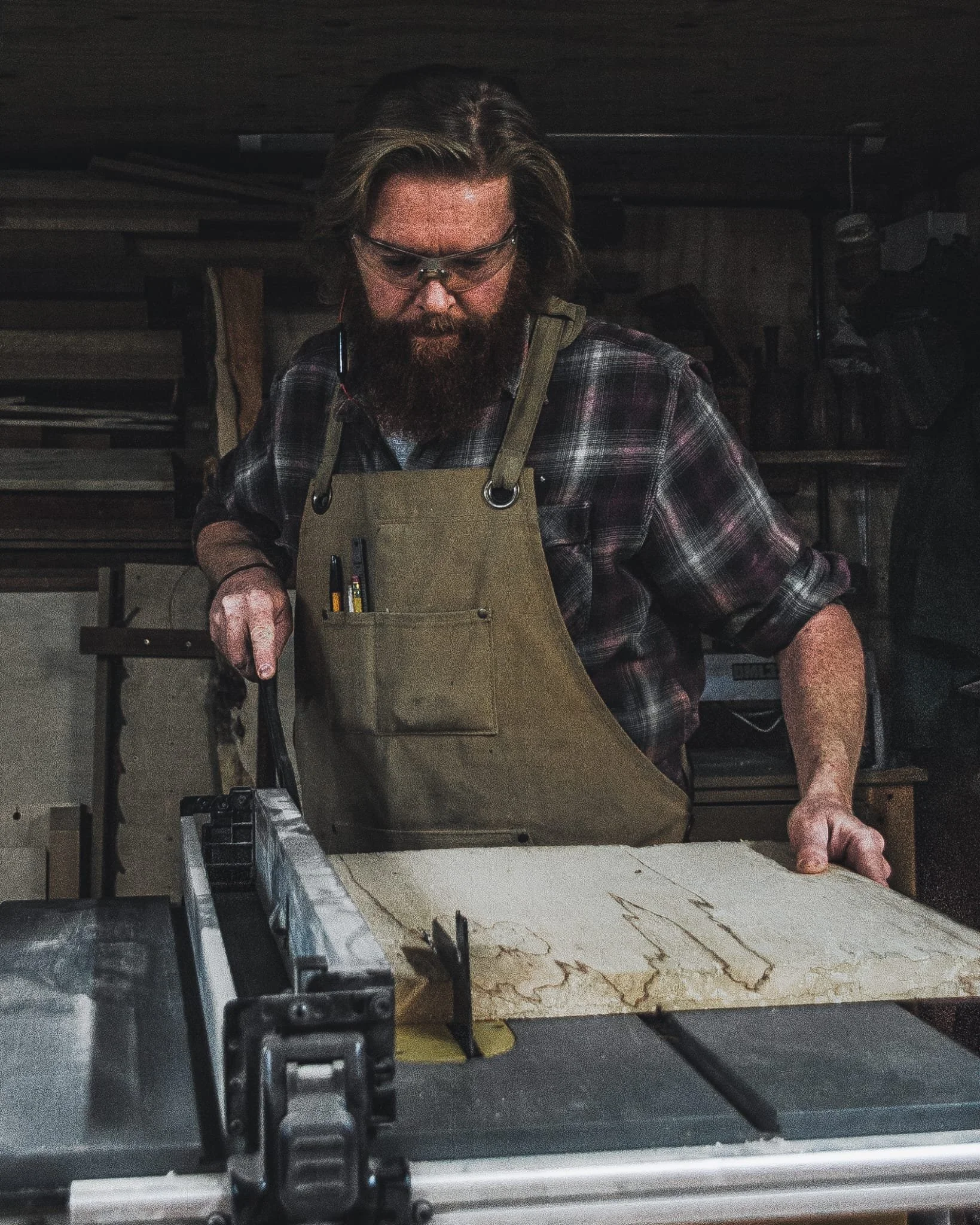 A man with glasses, a beard, and long hair, wearing a plaid shirt and beige apron, is using a table saw to cut a piece of wood in a woodworking shop.
