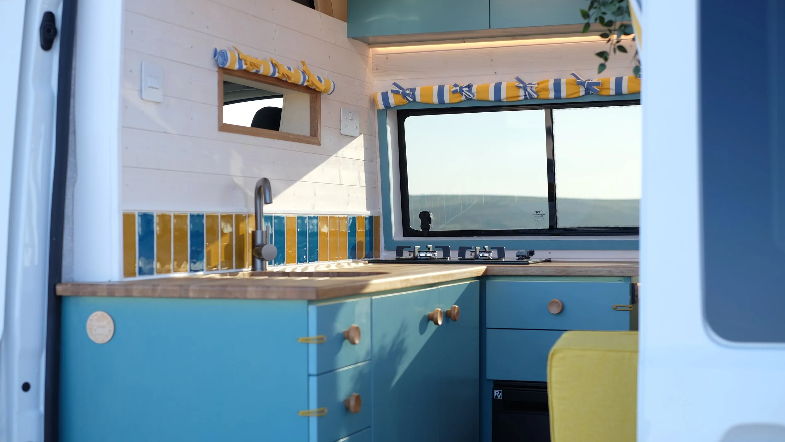 Inside view of a compact kitchen in a camper van with blue cabinets, a wooden countertop, a small sink, a window with striped valance, and a stove.