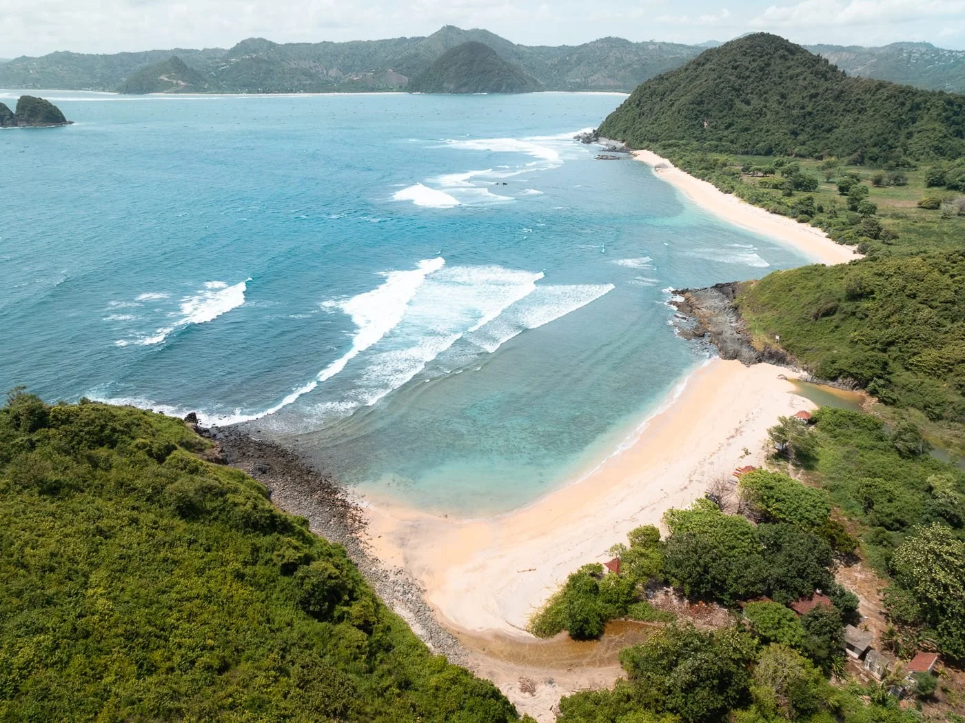 A scenic view of a beach with turquoise water, white sand, surrounded by lush green hills and distant mountains.