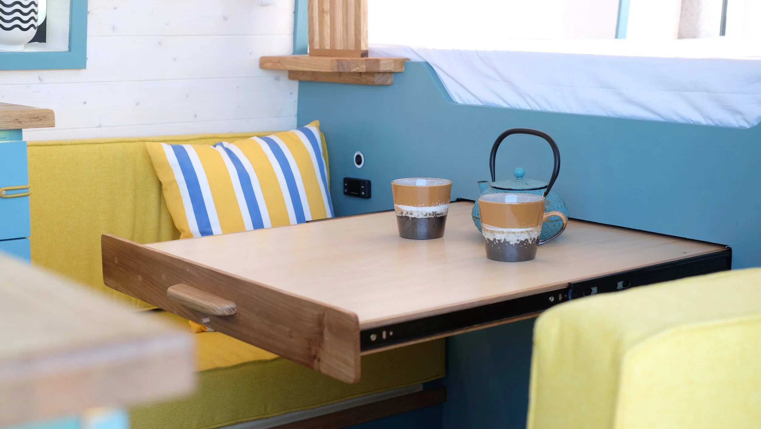 A cozy booth with a wooden table, yellow and striped pillows, a blue wall, and a teapot with two cups of coffee.