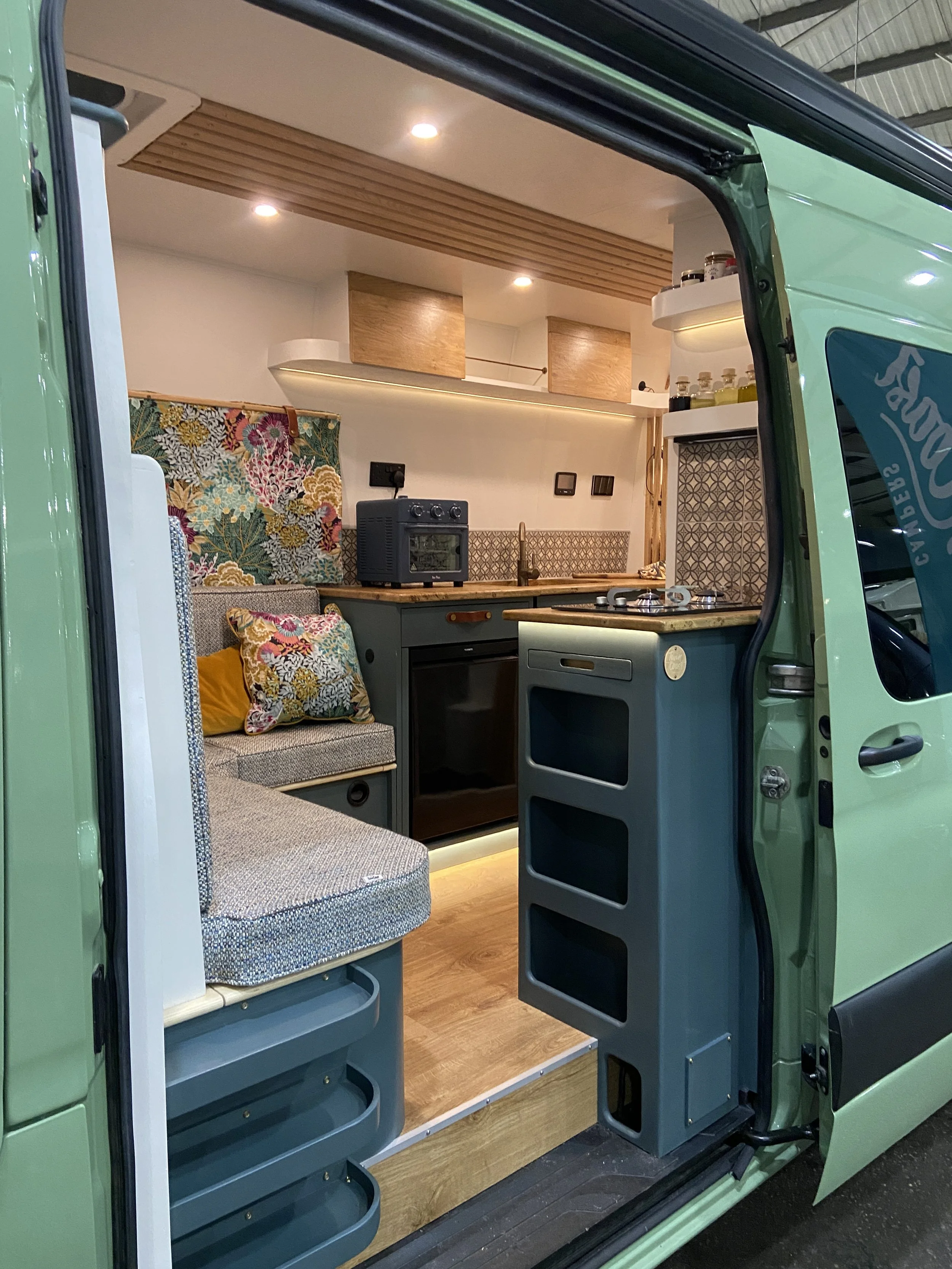 Interior of a compact camper van with a cozy kitchen and seating area, featuring colorful cushions, wooden cabinets, and decorative backsplash tiles, illuminated by built-in lighting.
