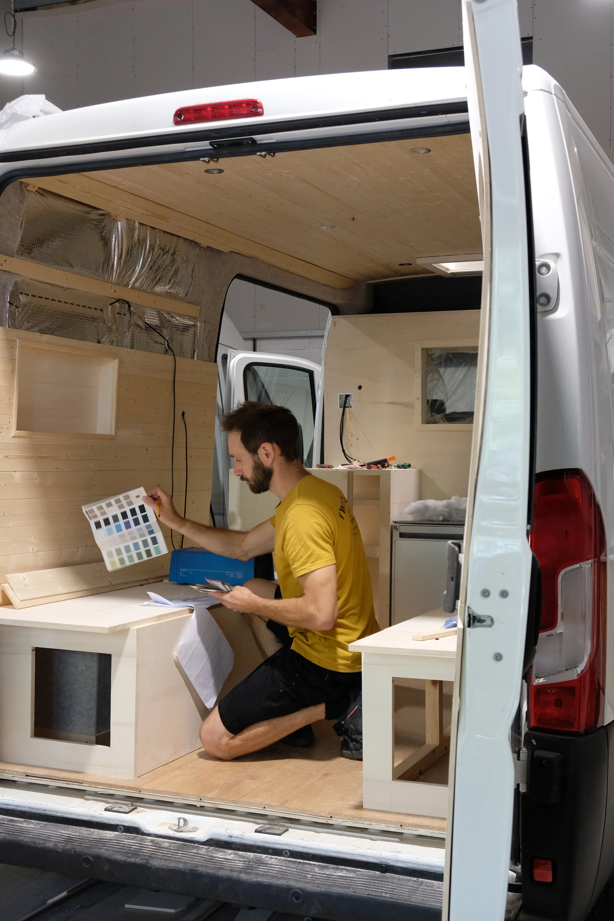 A man working inside a tiny home conversion van, examining color swatches and building with light-colored wood.