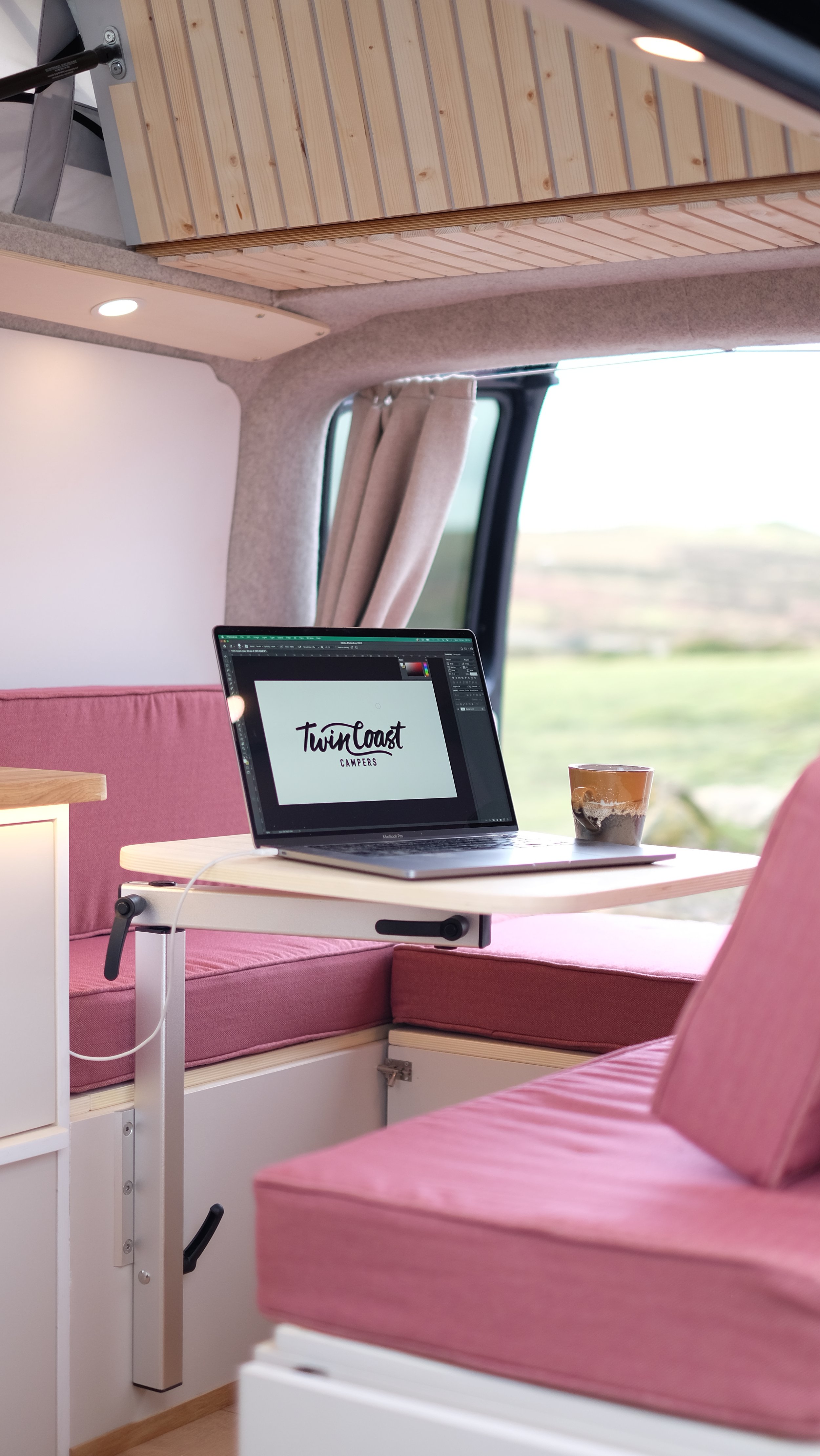 Inside a camper van with pink cushions, a small table with a laptop displaying the 'Twin Coast Campers' logo, and a glass of iced coffee. The van has wooden upper cabinets, beige curtains, and a window showing a landscape outside.