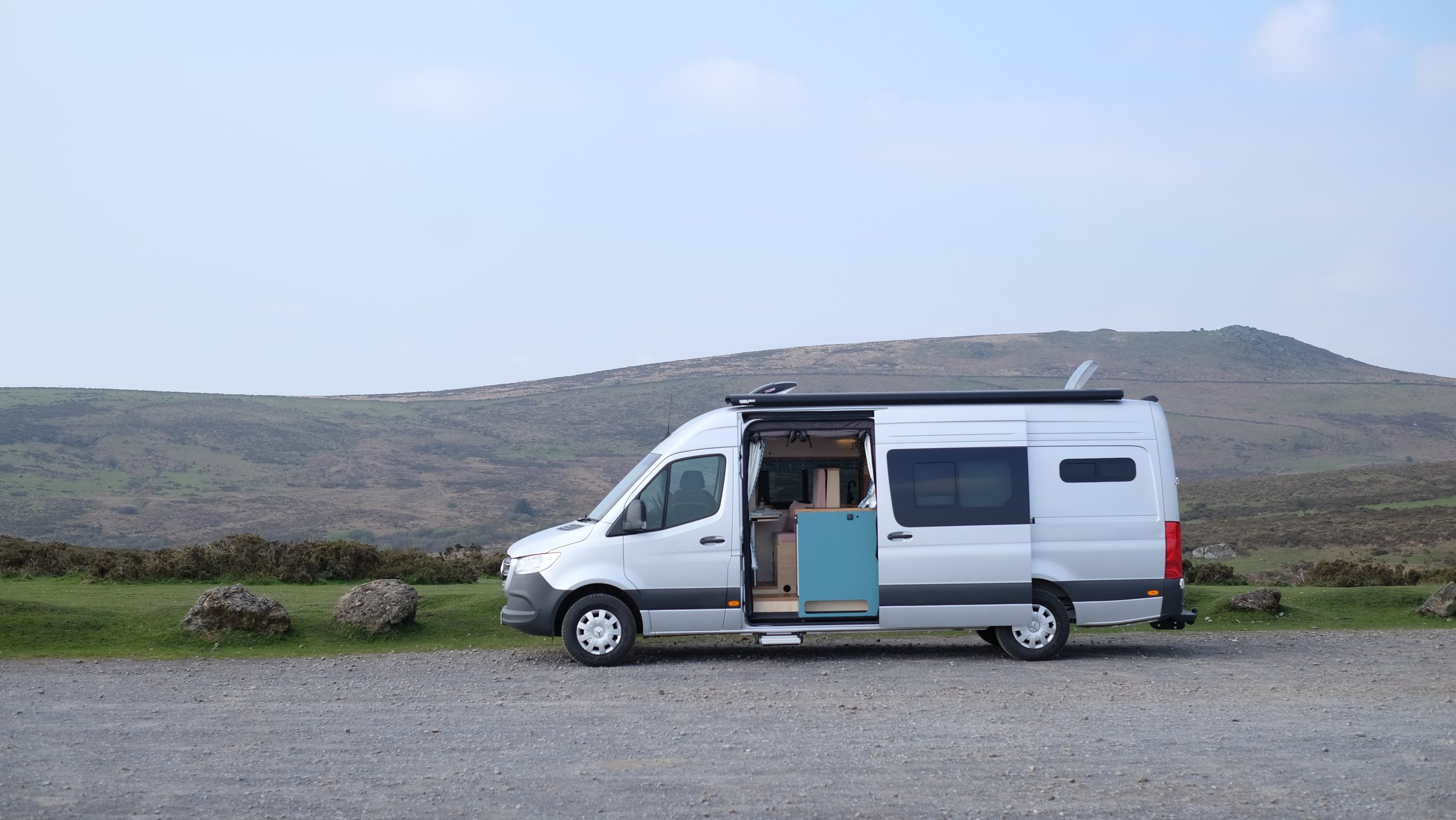 Silver camper van parked on a gravel area with open side door revealing the interior, set against a backdrop of hills and open sky.