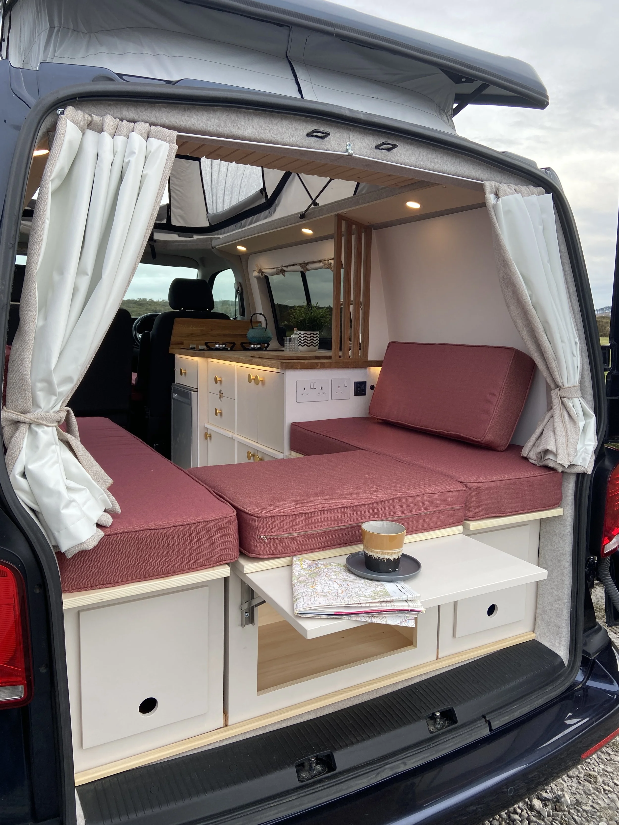 Interior of a camper van with pink cushions, white cabinetry, a small table with a cup and maps, curtains, and a view of the driver and passenger seats.