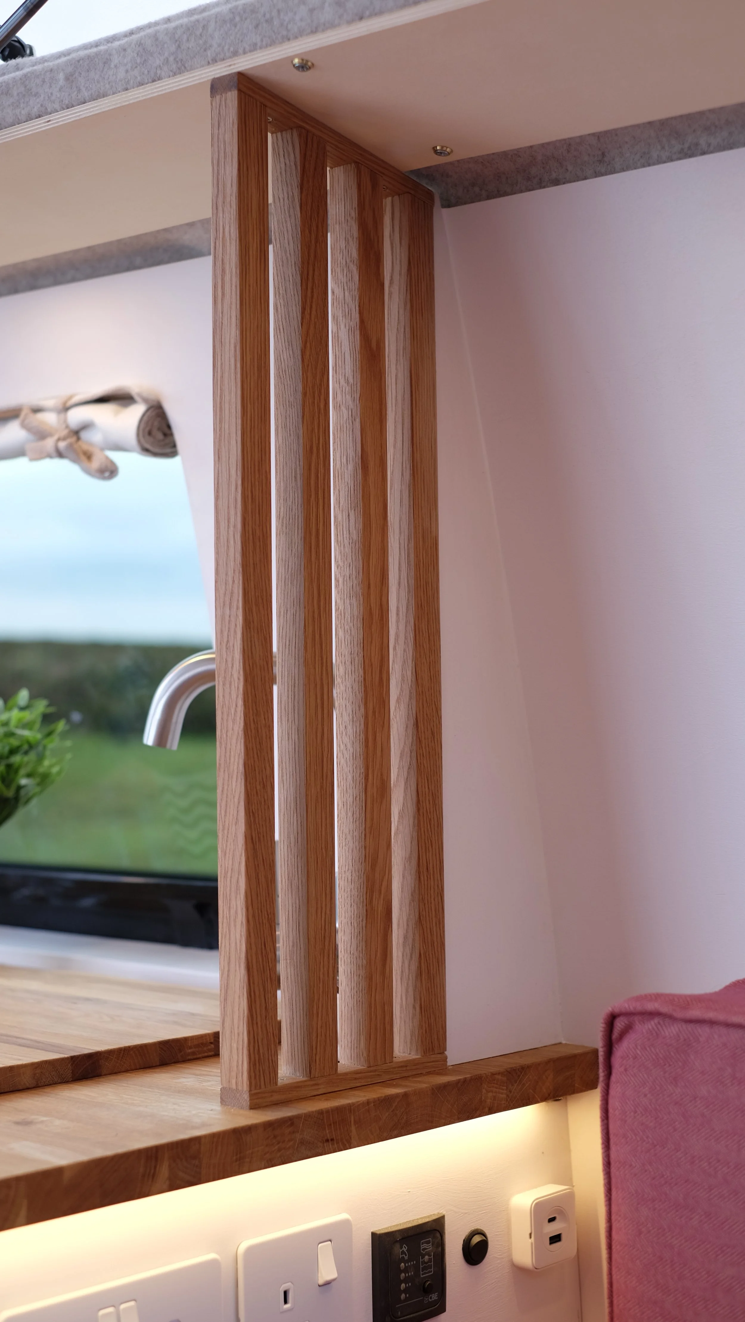 Close-up of a wooden placard with vertical slats, mounted on a countertop near a kitchen sink with a window showing outdoor greenery.