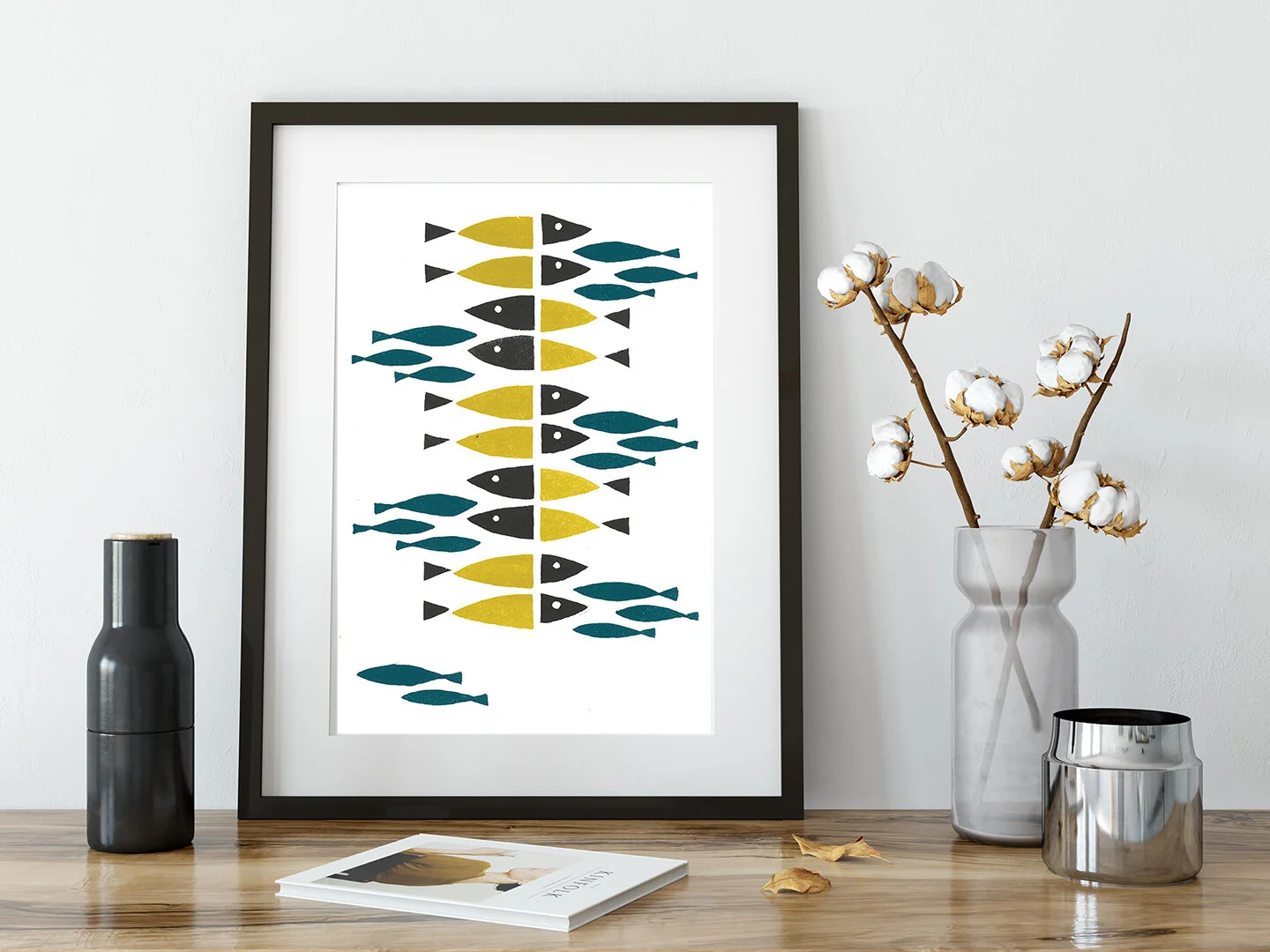 Framed print of abstract fish artwork displayed on a wood surface beside a cotton stem and modern decor items.