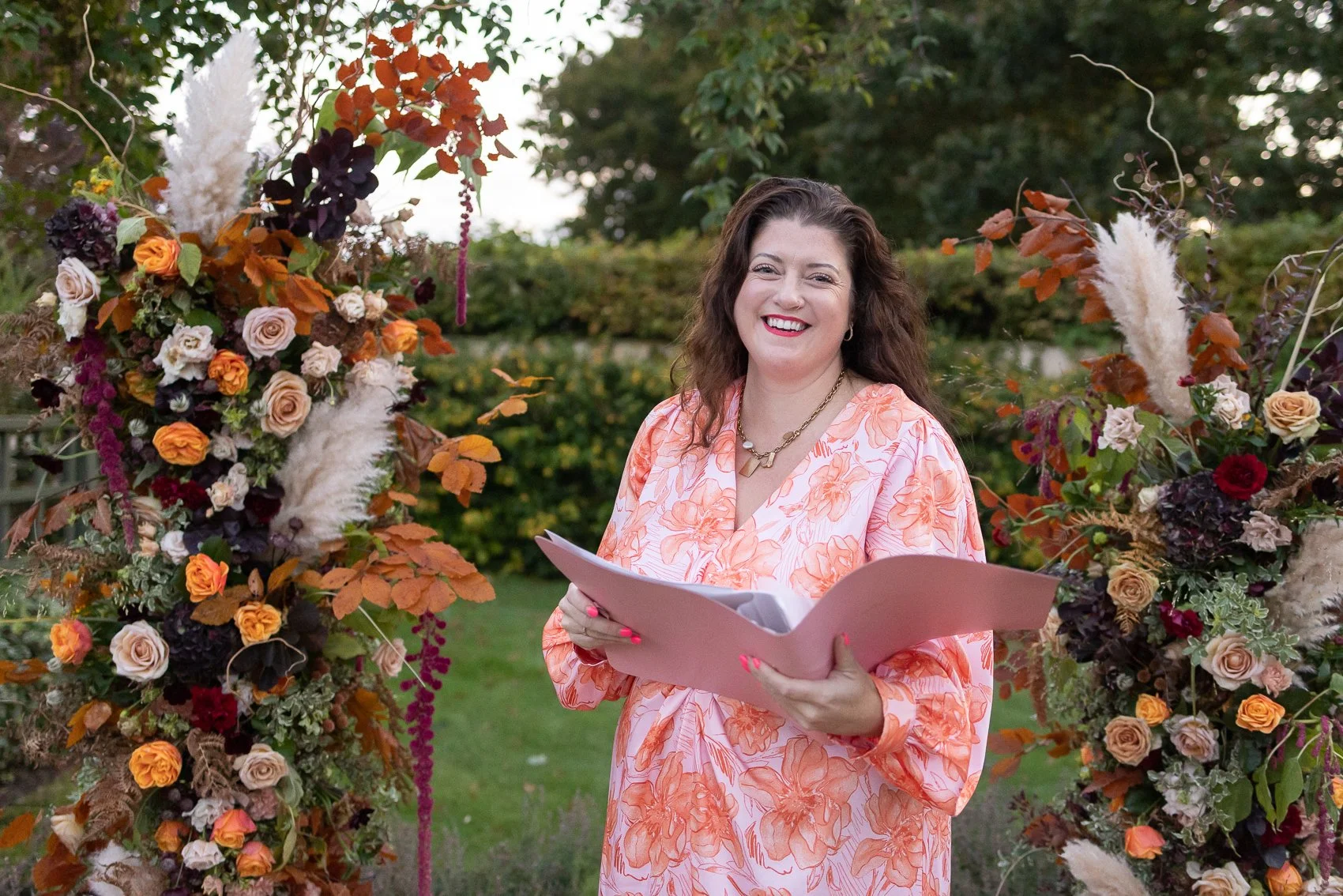 Portrait of Ashleigh Hitter, The Sunshine Celebrant, in an orange dress with a floral backdrop outside, conducting a wedding with a pink folder in her arms