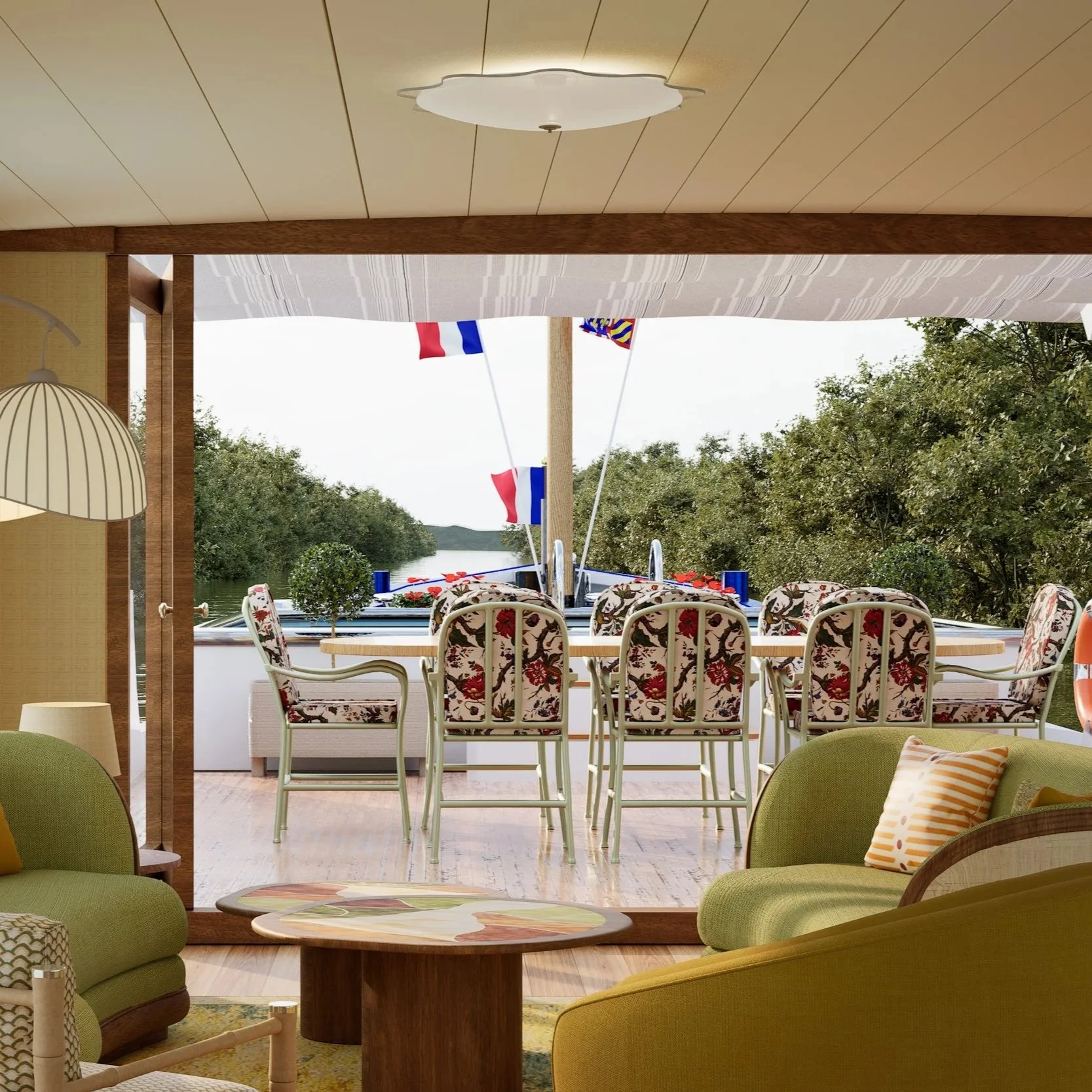 A cozy indoor space opens up to a balcony overlooking a scenic waterfront with trees and a cloudy sky. Inside, there are green chairs with patterned pillows, a round wooden coffee table, and seating around a dining table with floral chairs. The porch has flags, including the French flag, displayed above the water.