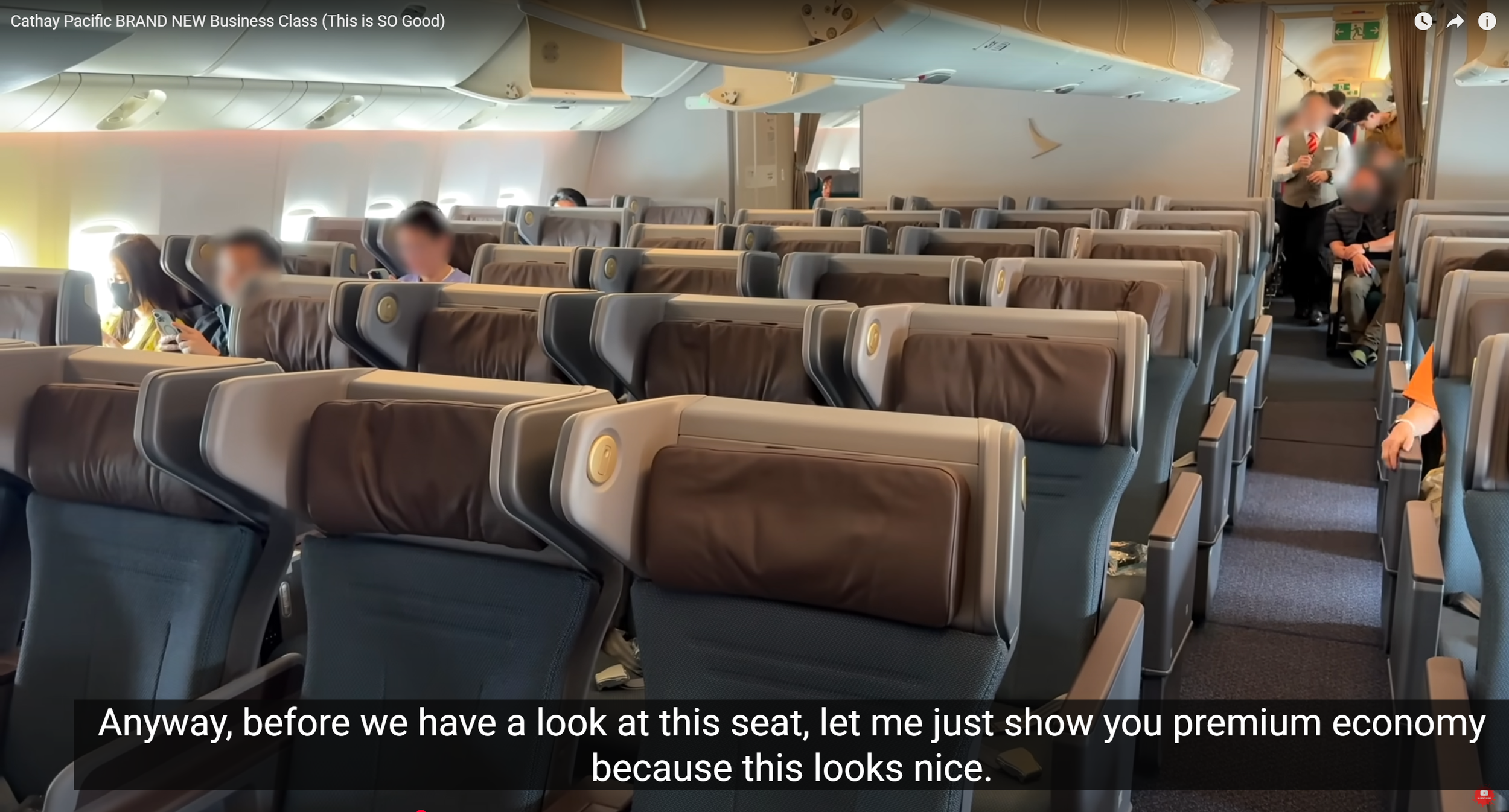 Interior of an airplane cabin with several rows of seats, some passengers sitting and some standing near the aisle. The seats have brown leather headrests, and a few passengers are using electronic devices. Overhead compartments are closed, and a ste