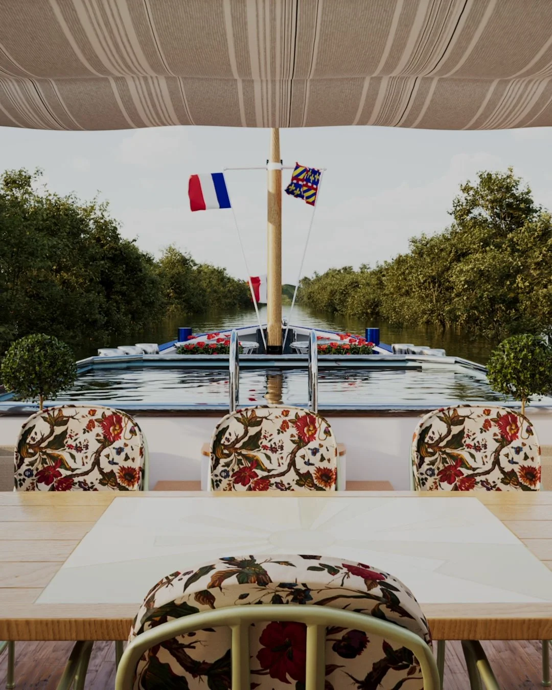 A boat decorated with floral arrangements and flags is seen from a dining area with floral chairs, on a river lined with trees.