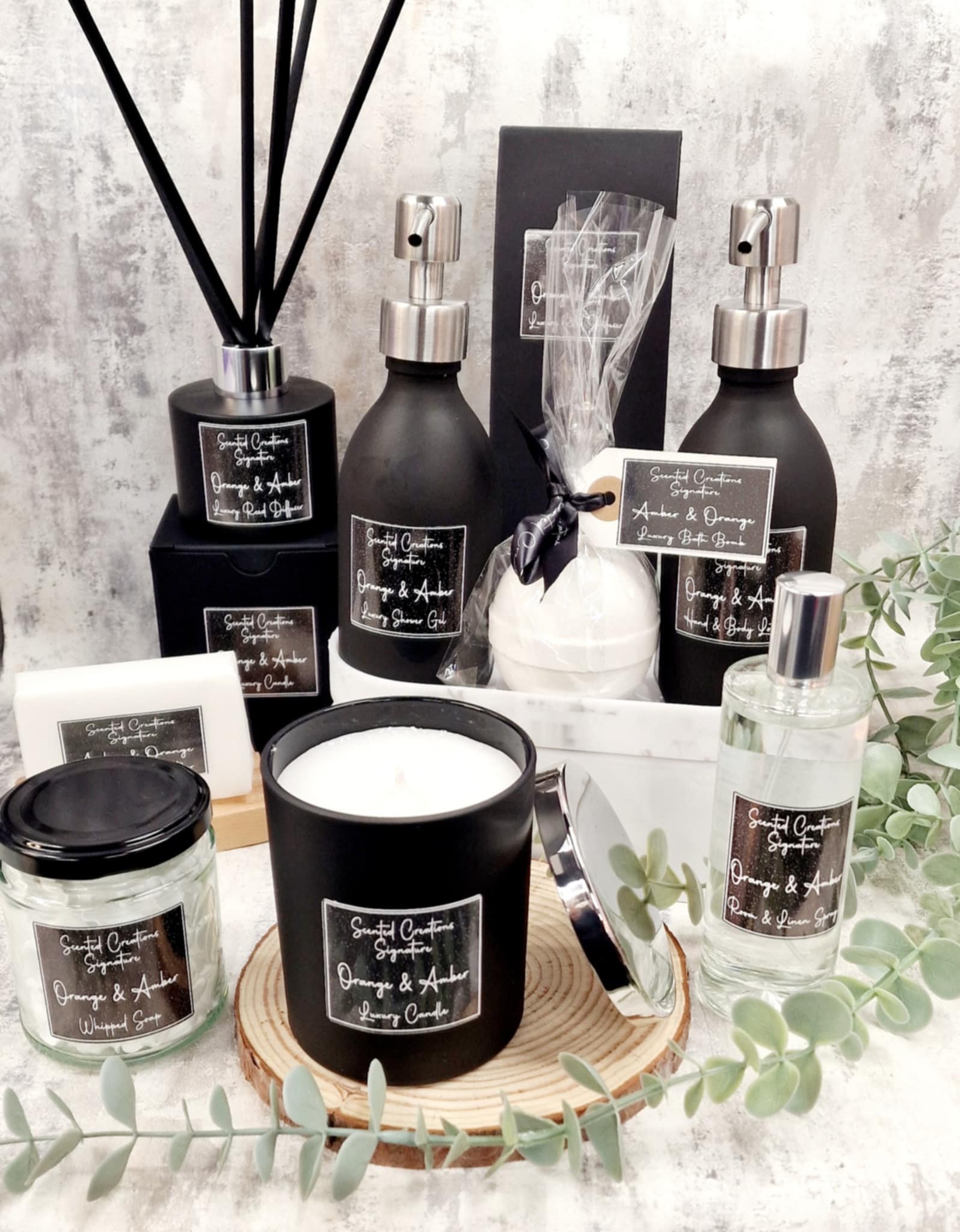 Collection of orange and amber scented products, including candles, reed diffuser, lotions, and sprays, displayed on a rustic wooden surface with foliage.