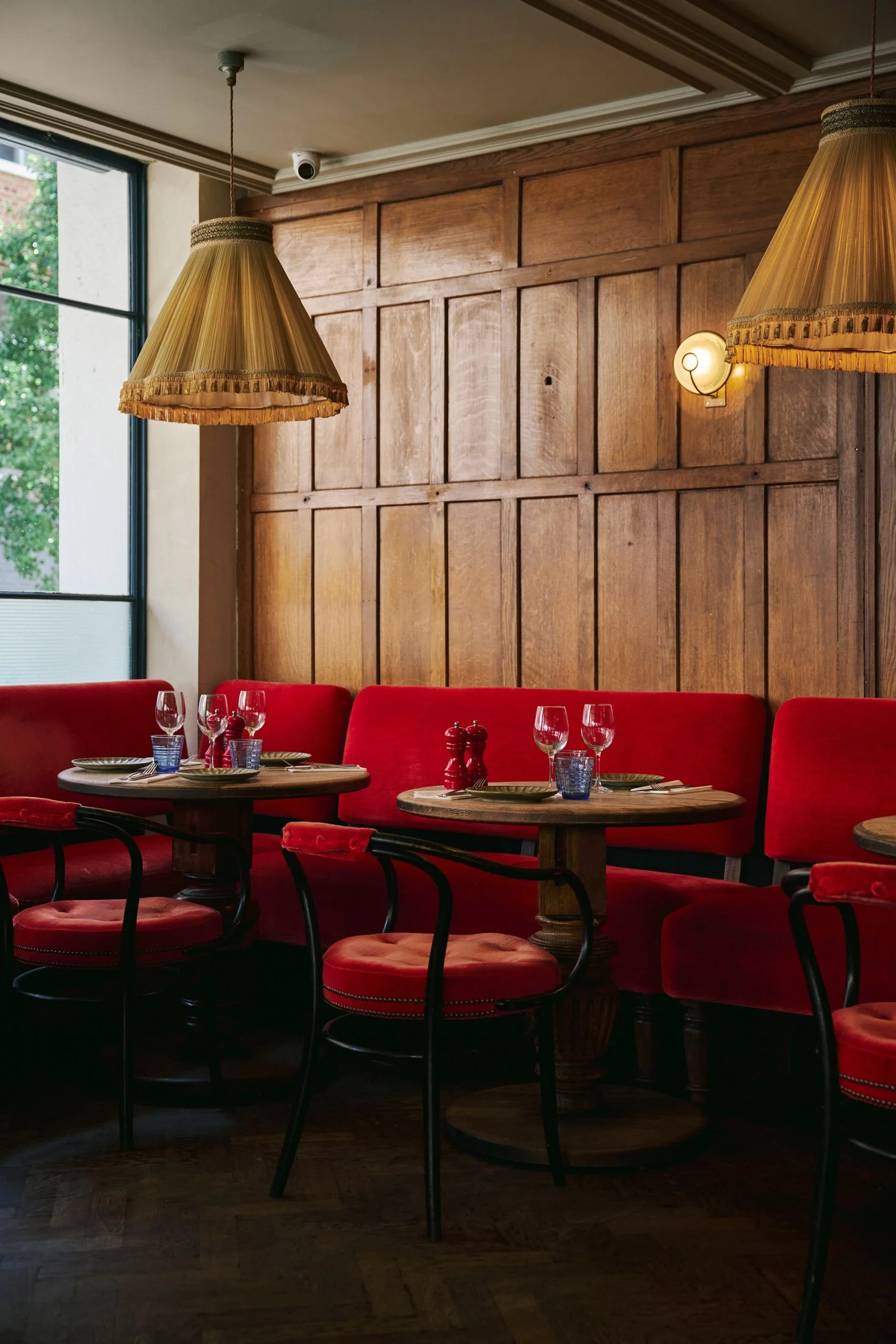 Cozy restaurant interior with red velvet seating, round wooden tables set with glassware and plates, against a paneled wood wall. Interior photographer London.