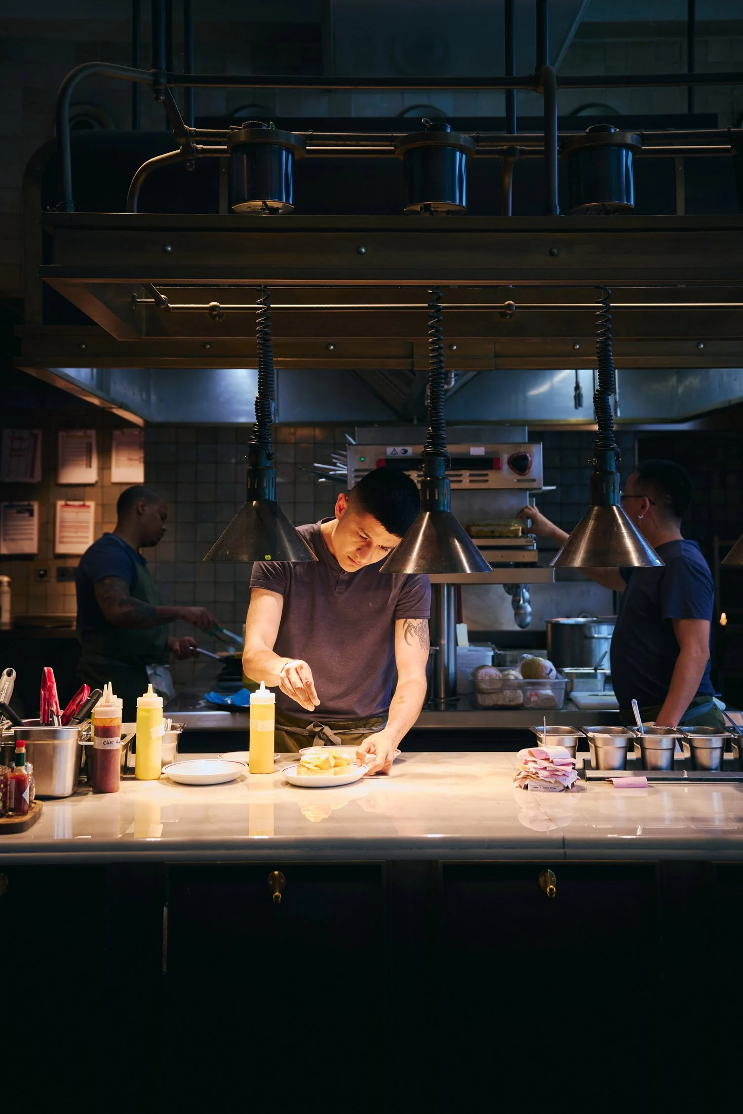 Chef preparing a dish in a restaurant kitchen under hanging lights, with other kitchen staff working in the background. Lifestyle photographer London.