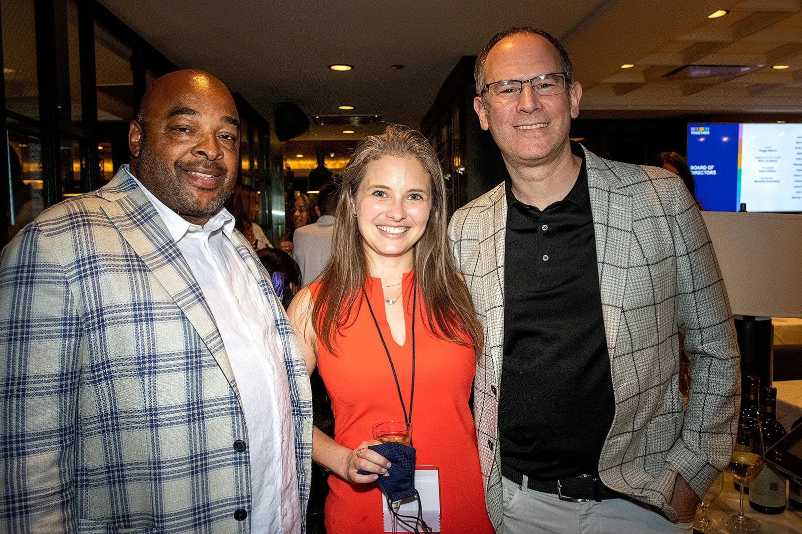 Carl West, Lauren Schrero, and Rick Jacobson pose in the Steak 48 venue. To the left, Carl wears a beige and blue plaid suit jacket. In the center Lauren wears a dark orange dress and holds a drink in her right hand. To the right, Rick, who is TNP's