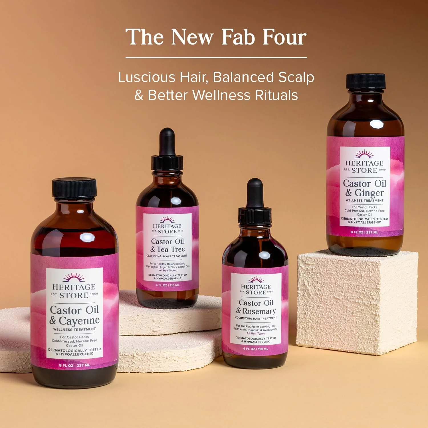 Four bottles of Heritage Store castor oil and scalp treatment products displayed on beige blocks against a light brown background. The bottles have pink labels with black text and black caps, some with dropper tops. The text above the bottles reads, "The New Fab Four: Luscious Hair, Balanced Scalp & Better Wellness Rituals."