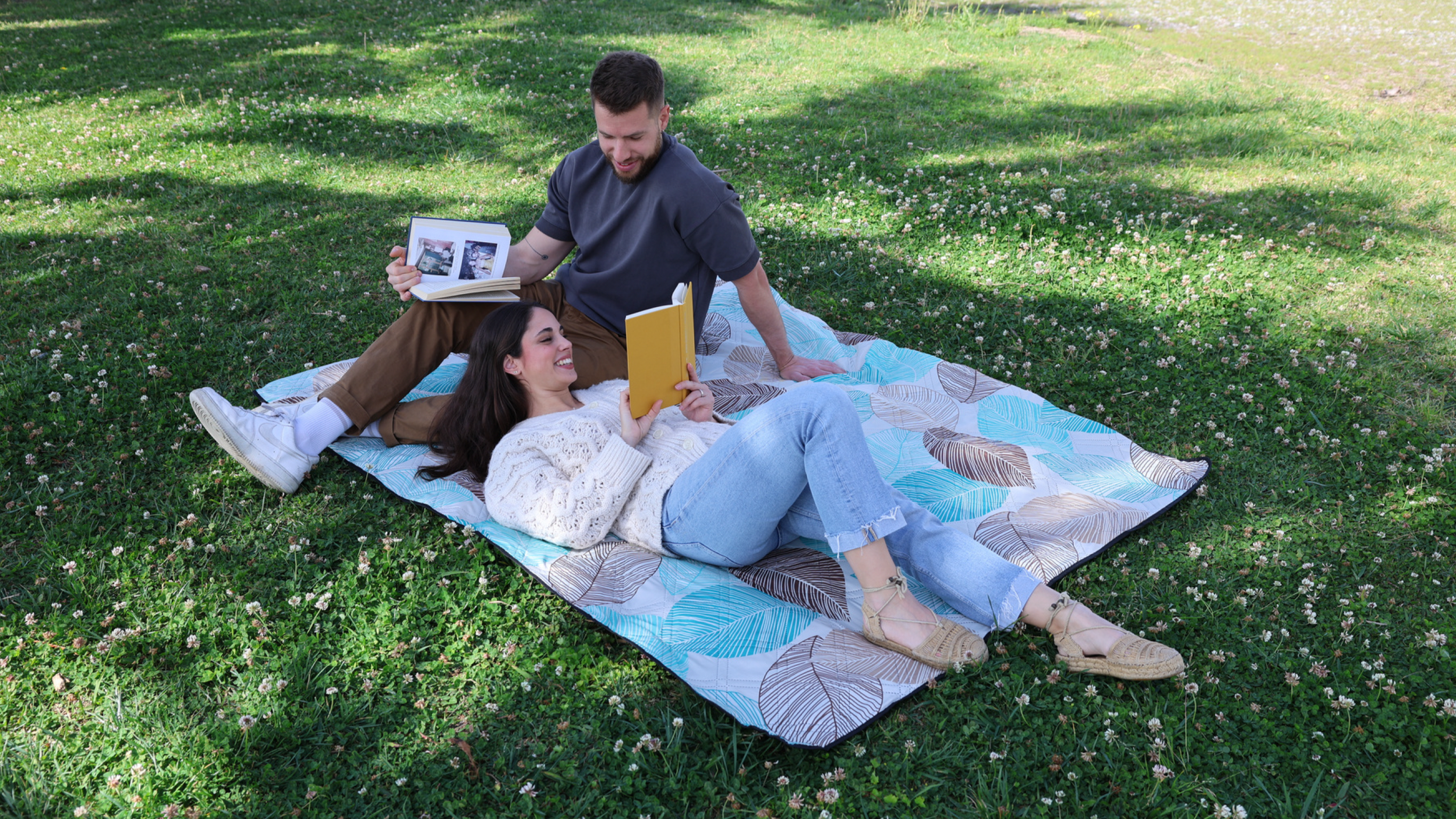 A young woman and man relaxing on a blanket in a grassy park, reading books and smiling at each other.