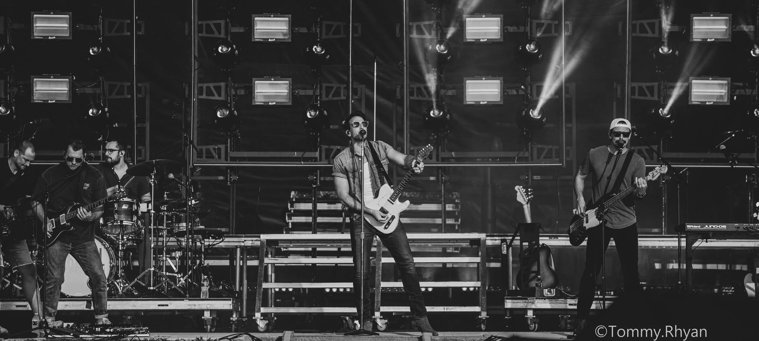 Black and white photo of a band performing on stage. Musicians play guitars and drums, while stage lights illuminate behind them. The scene conveys energy and live music atmosphere.
