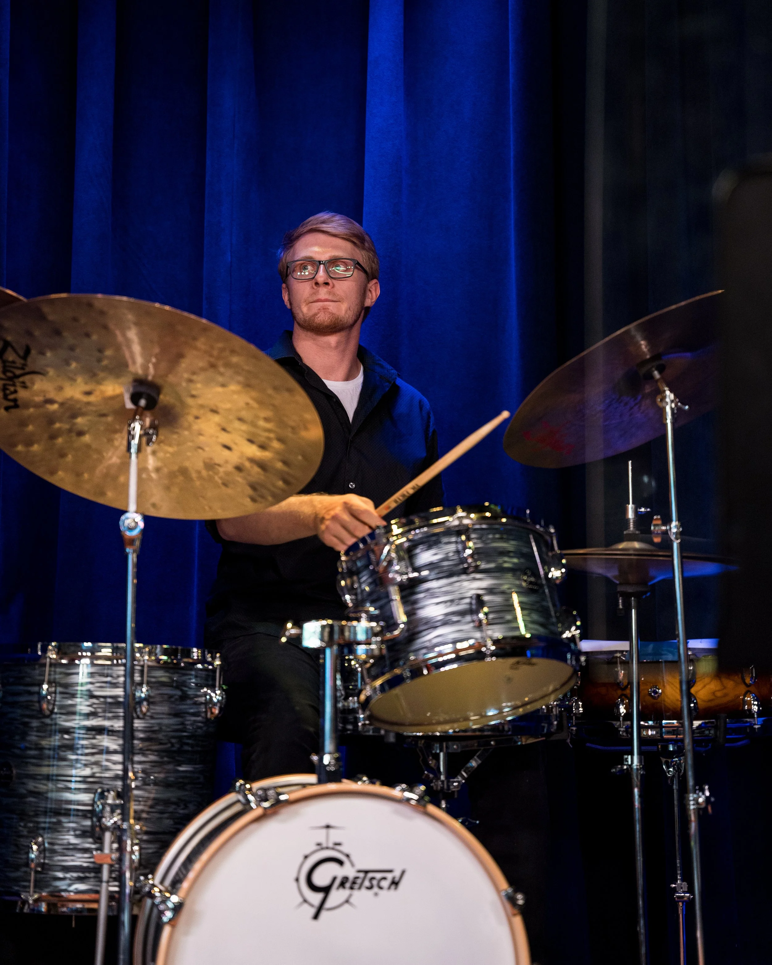 Drummer playing a Gretsch drum set on stage with blue curtains in the background.