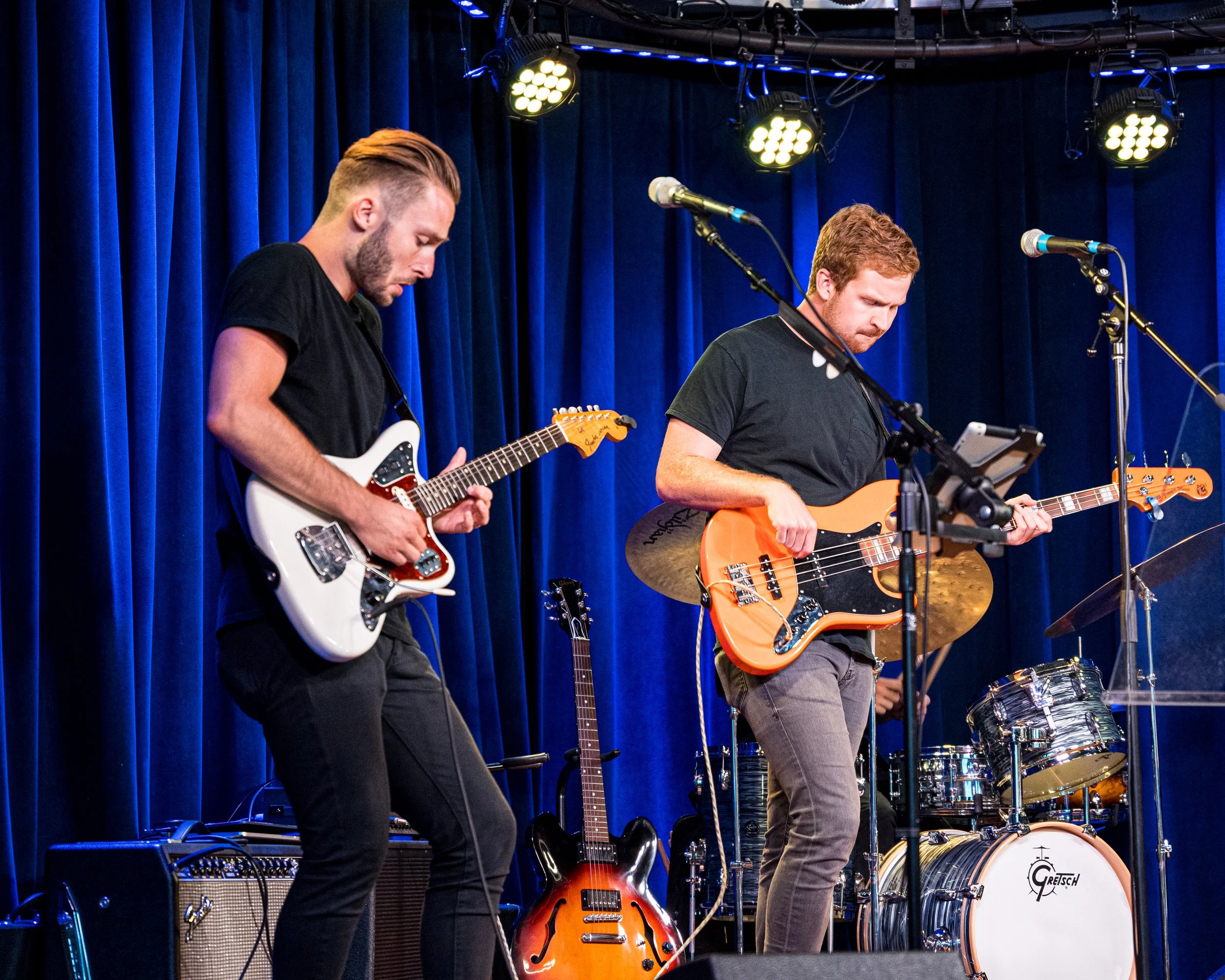 Two male musicians performing on stage with electric guitars and blue curtains in the background.