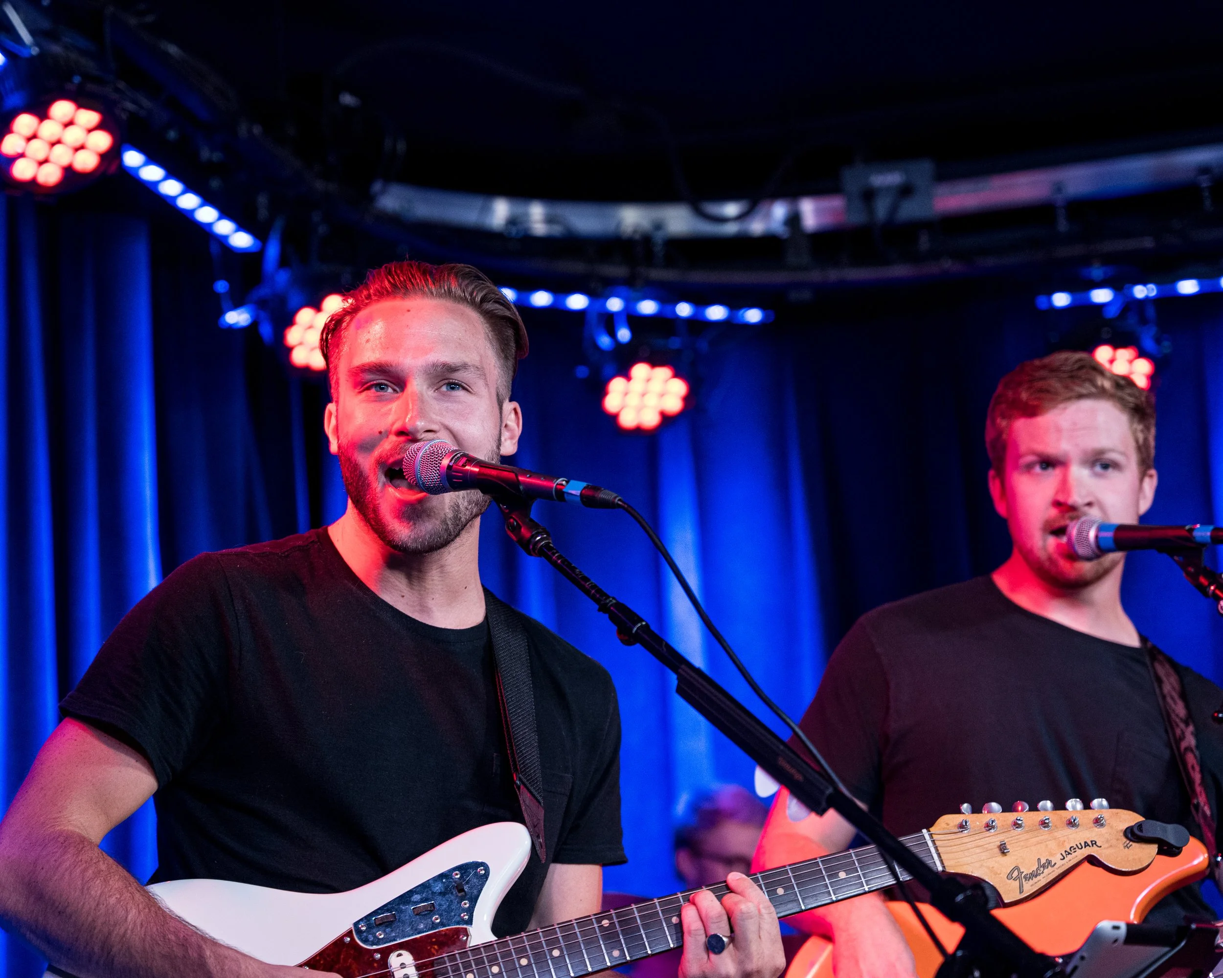 Two musicians performing on stage with guitars and microphones, blue and red stage lighting.