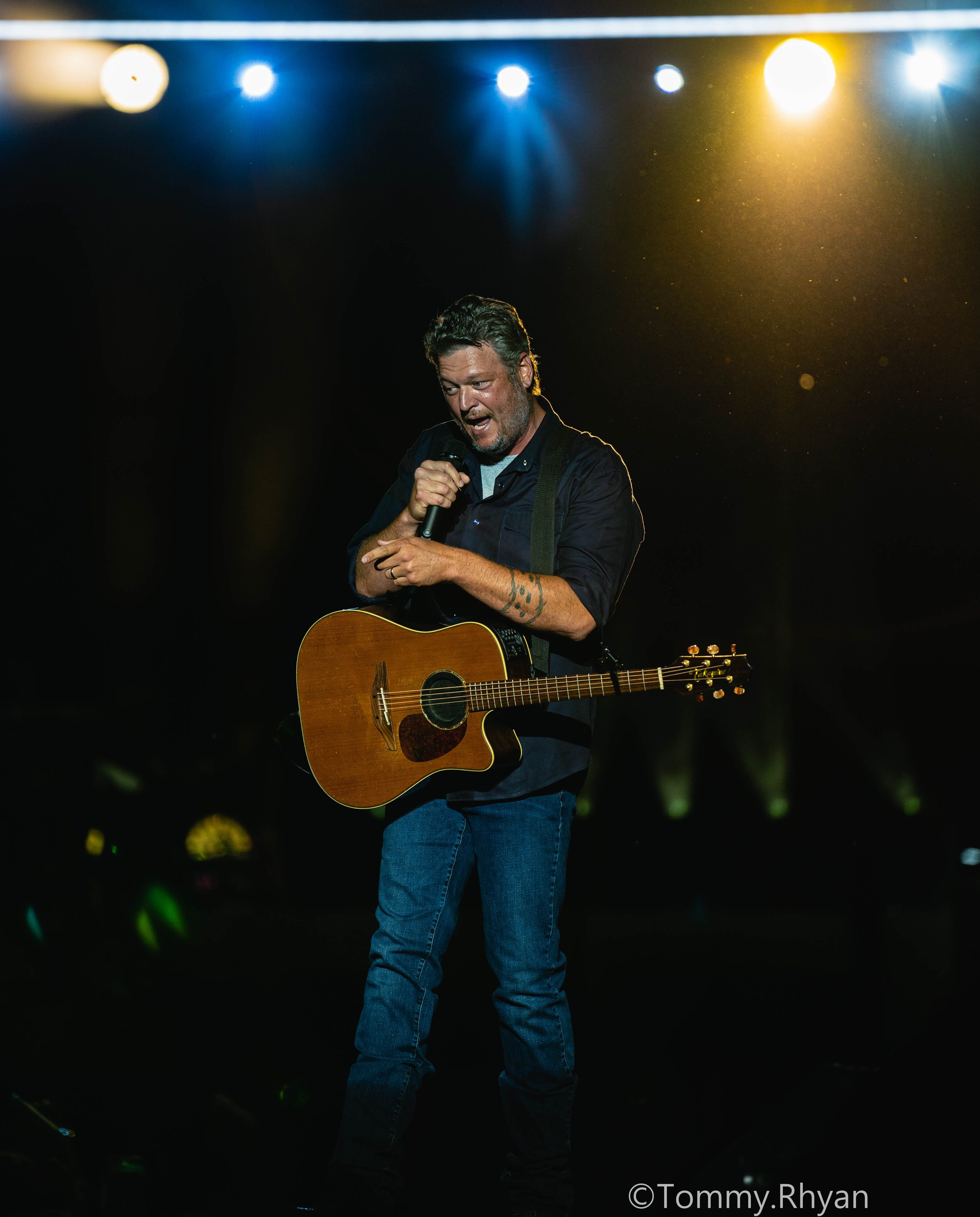Man holding a microphone and guitar, performing on stage with bright lights in the background.