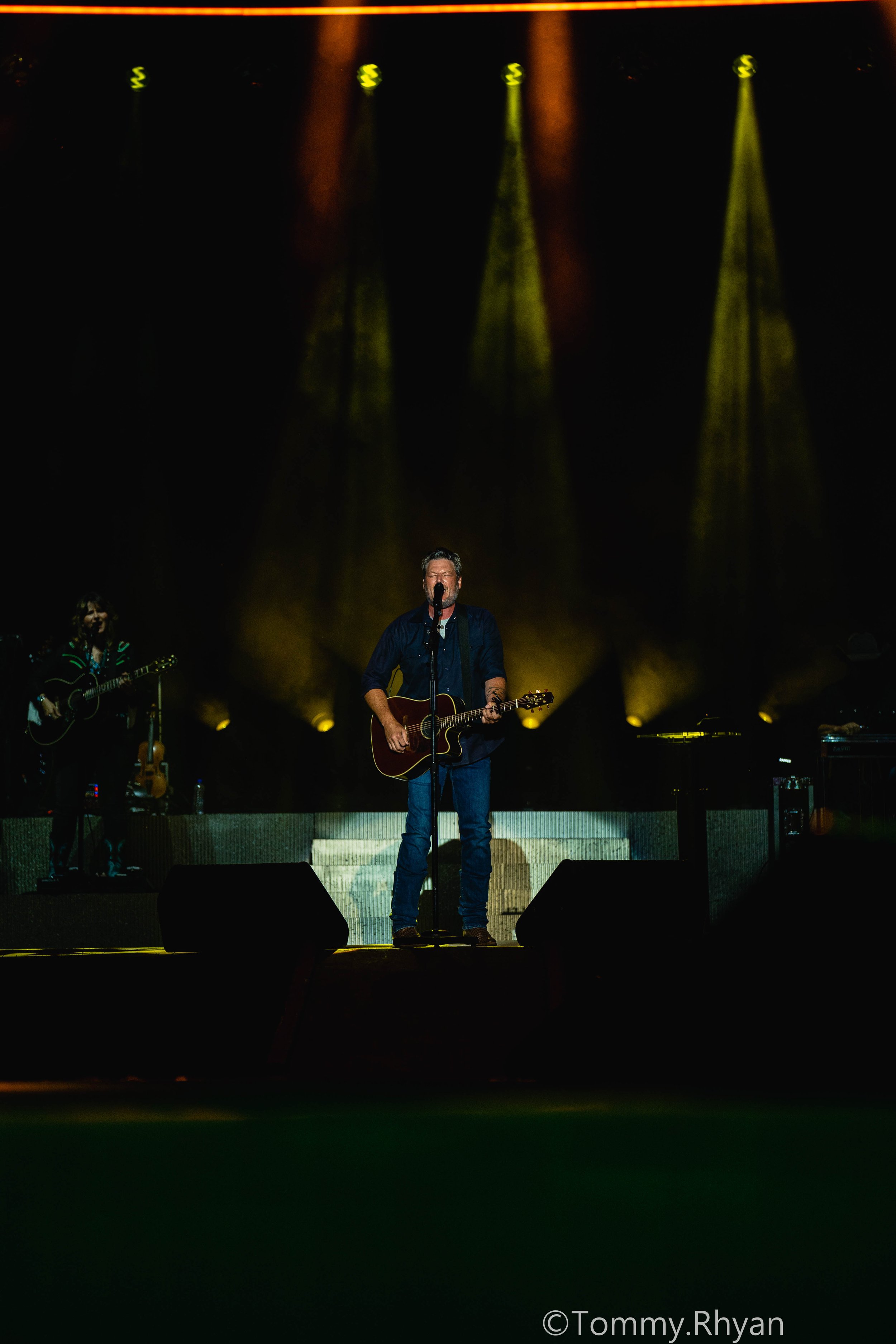 Country musician performing on stage with a guitar, illuminated by yellow and orange stage lights, accompanied by another musician in the background.
