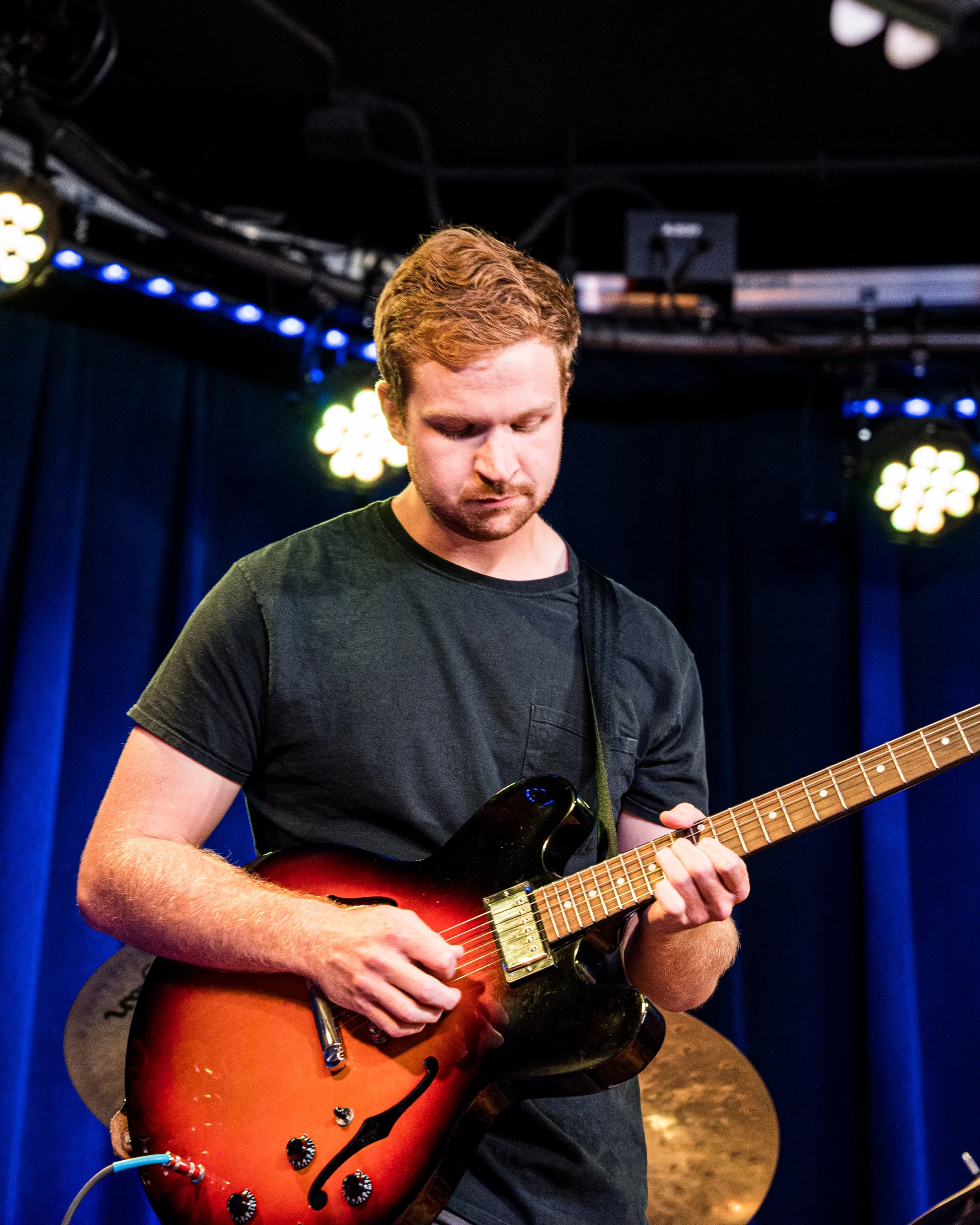 A man playing an electric guitar on stage with blue curtains and stage lights in the background.