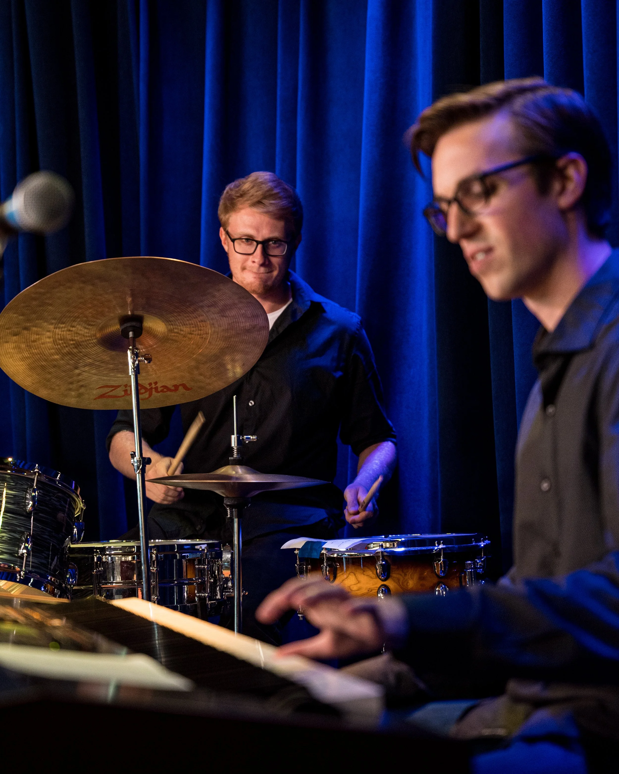 Musicians performing on stage, one playing drums and another playing piano with blue curtains in the background.