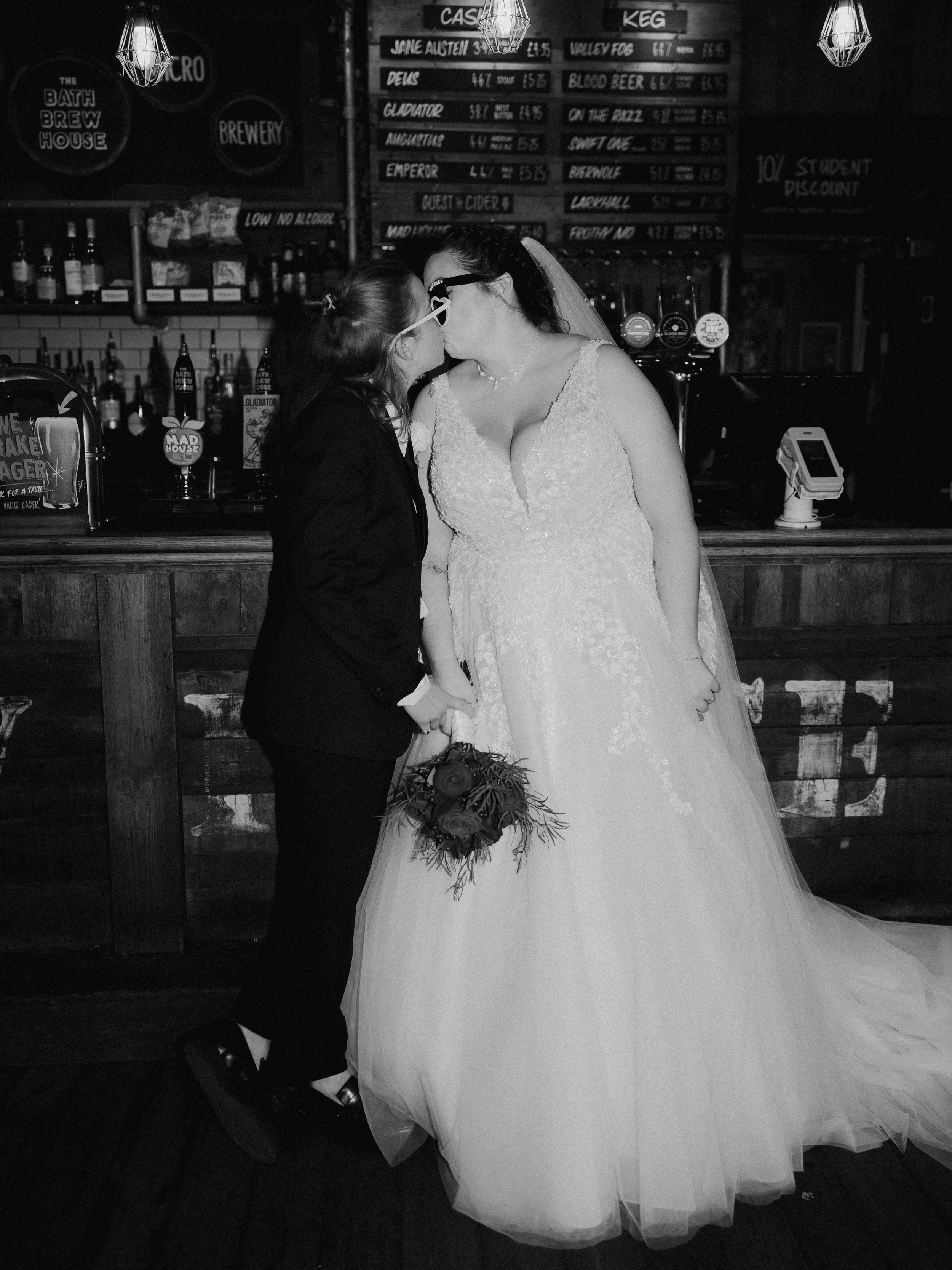 A black and white editorial image of two brides posing against a pub bar, with heart shaped wedding sunglasses on.