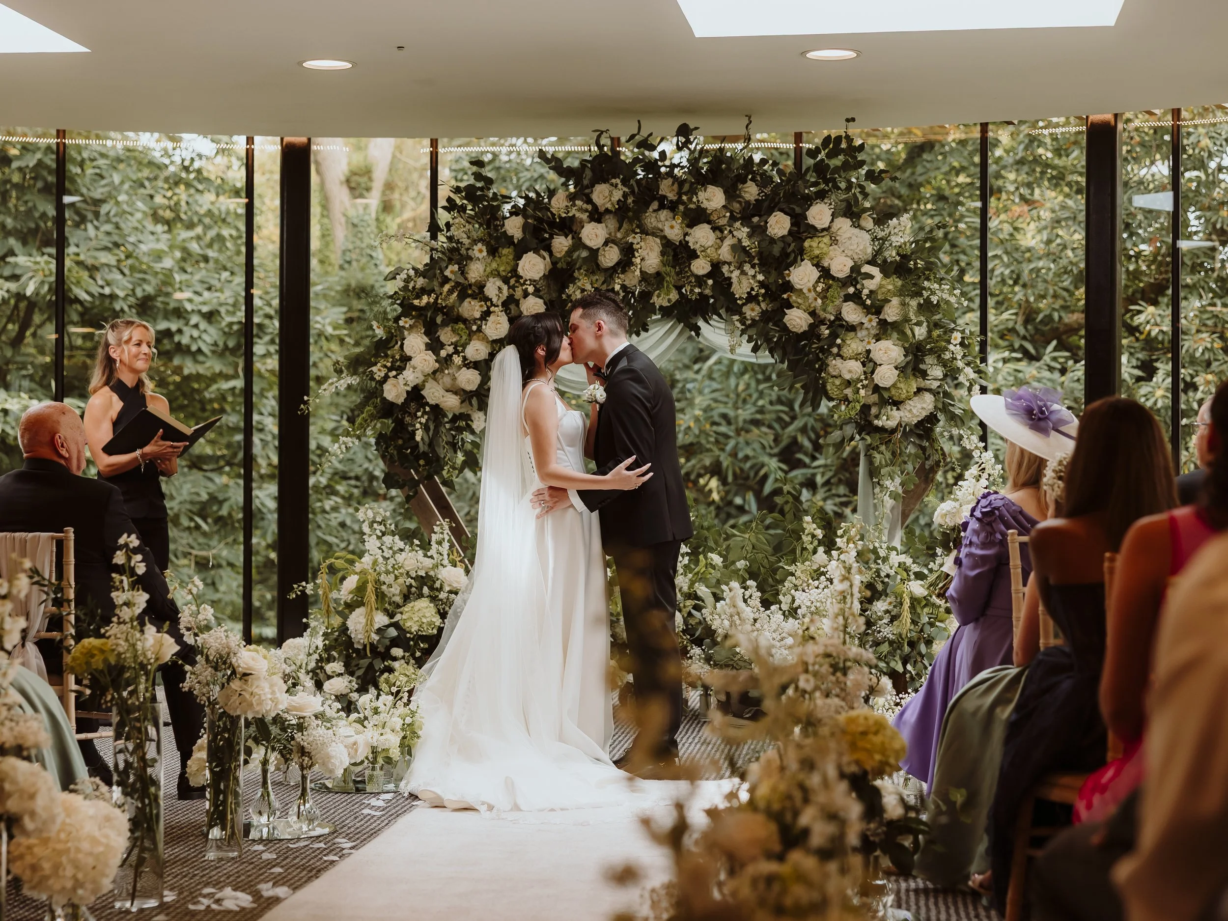 Intimate indoor wedding ceremony with floral arch, photographed in a relaxed, documentary style