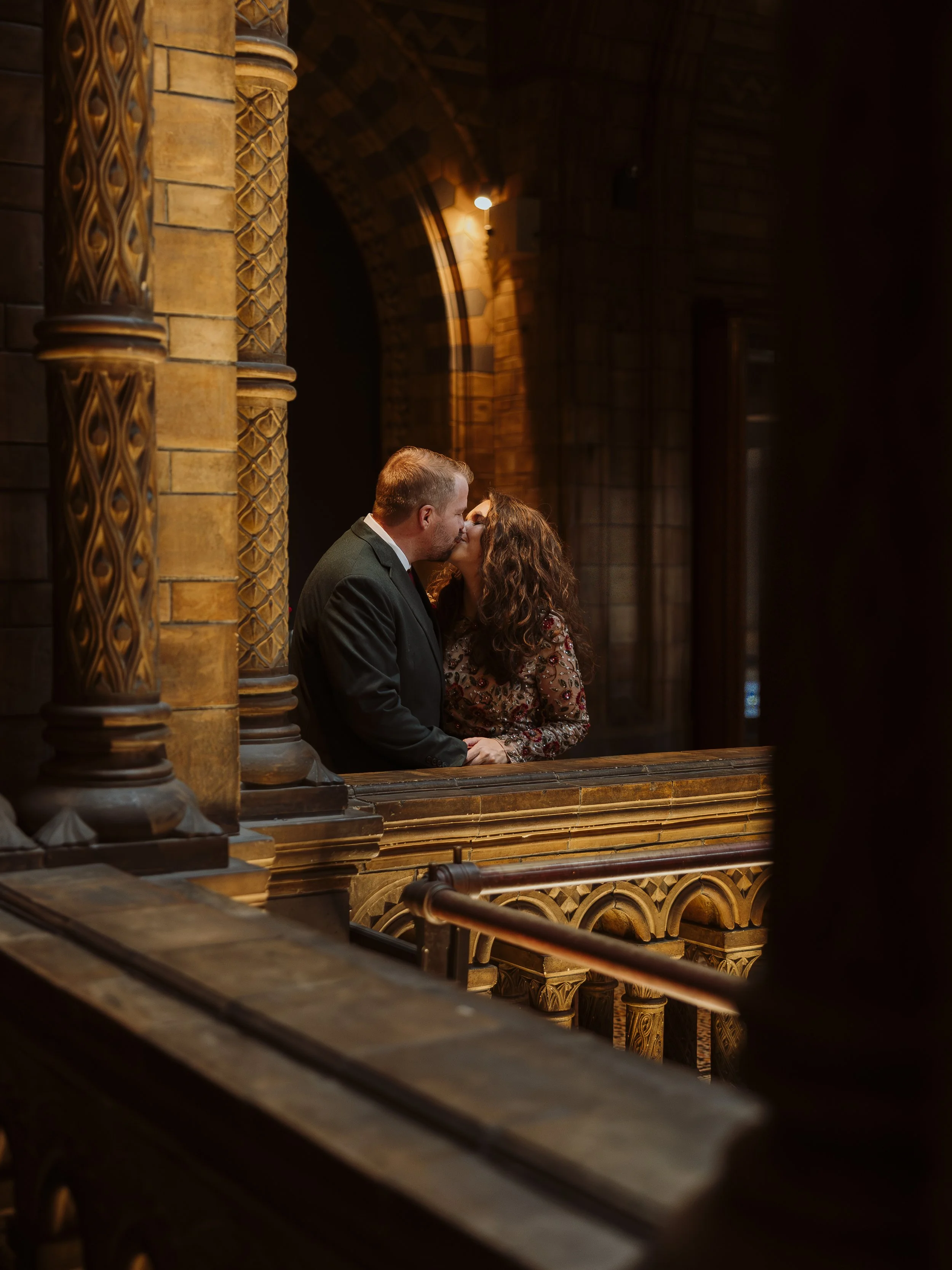 Bride and groom holding hands inside the Natural History Museum in London, surrounded by historic stone arches.