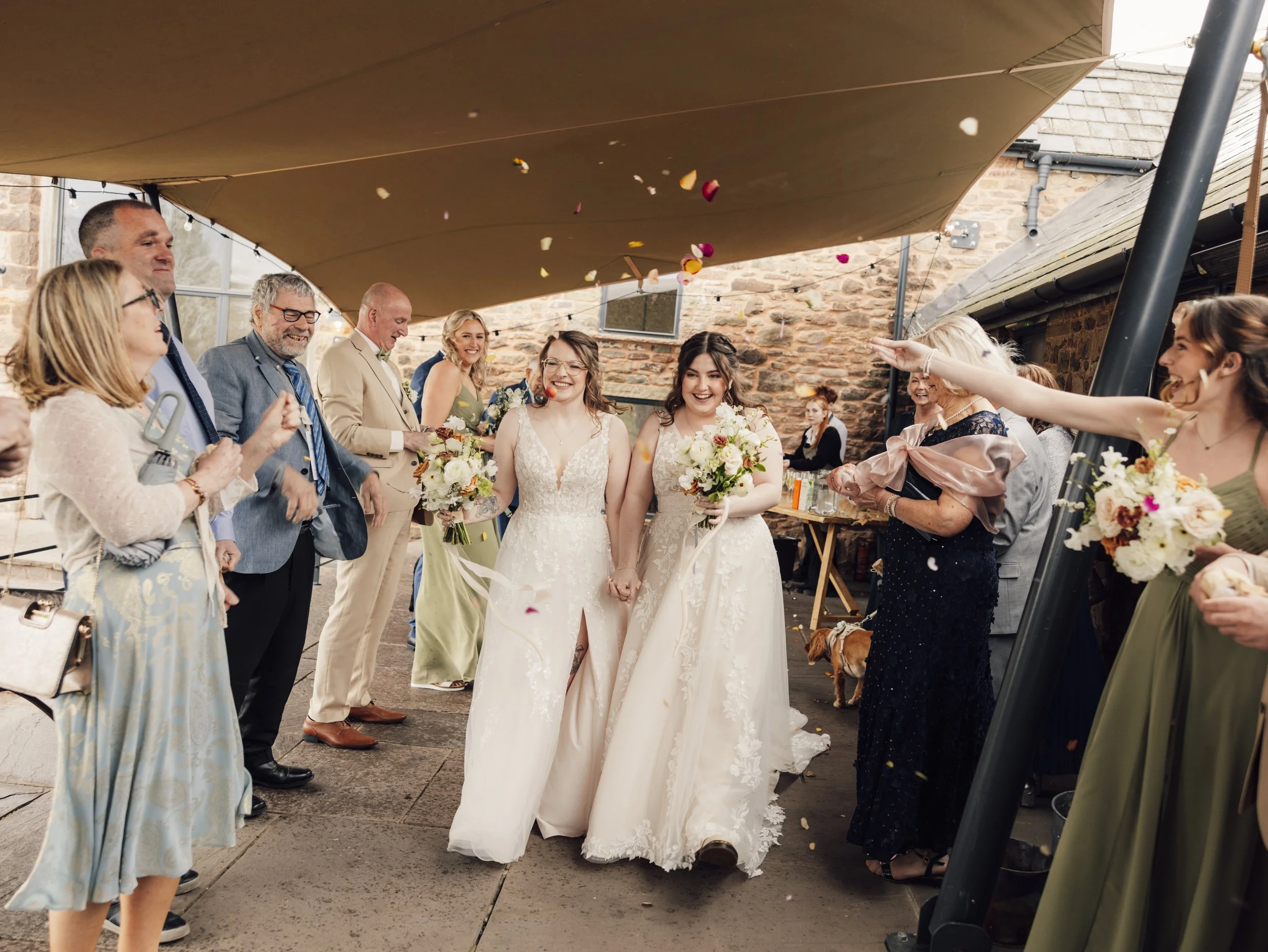Bride and bride walking hand-in-hand through a confetti tunnel at their Llantilio House wedding, surrounded by smiling guests under an outdoor canopy.