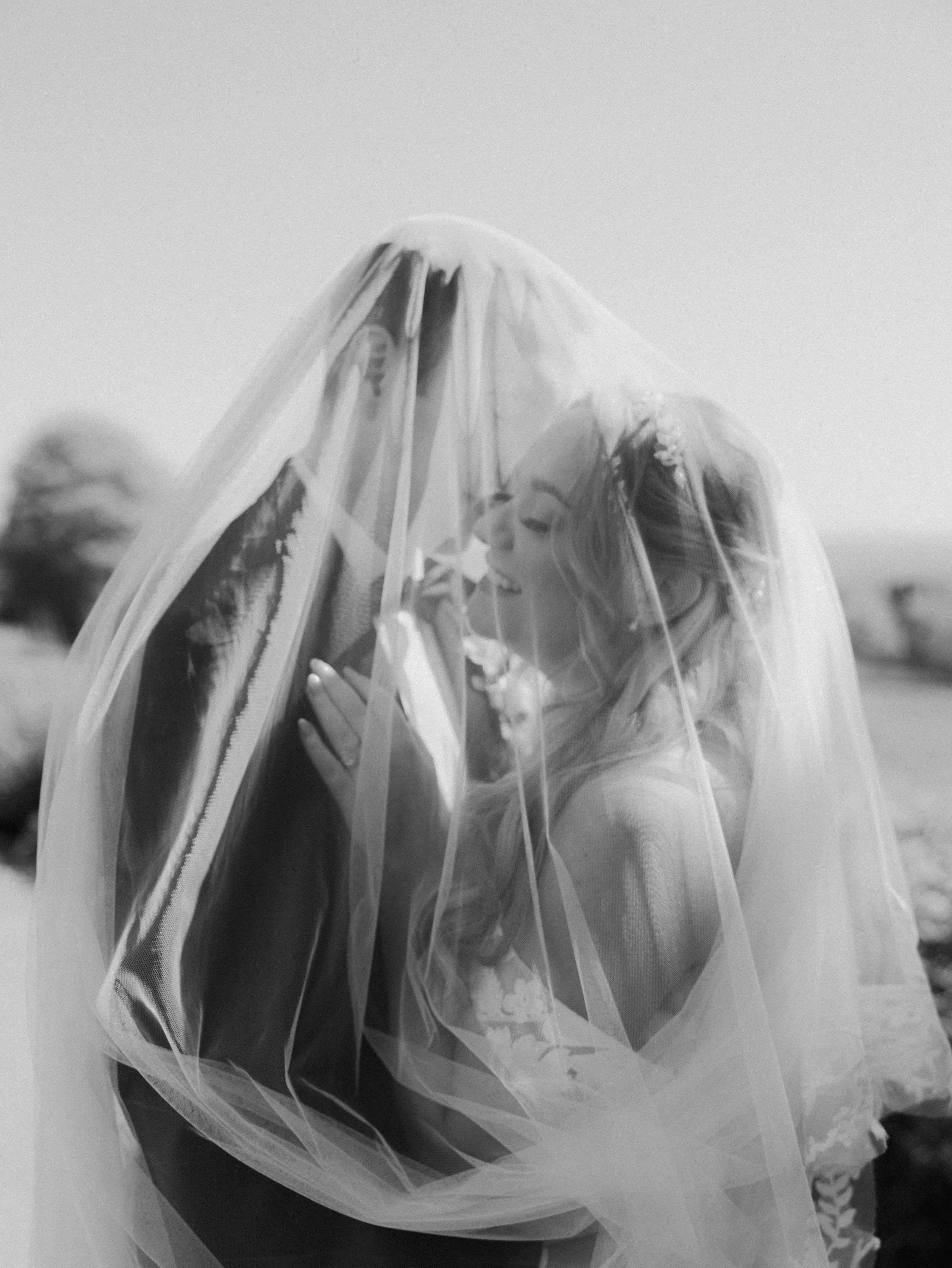A bride and groom embracing closely under the brides veil, captured in black and white.