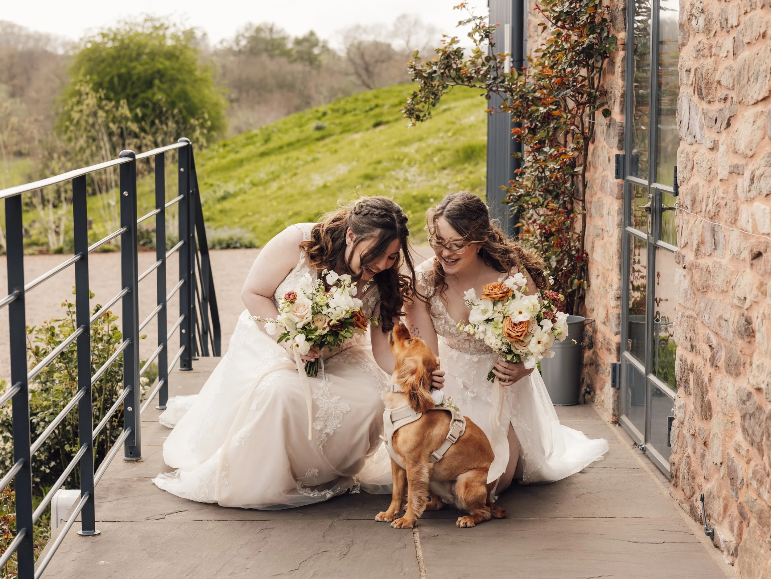Two brides in lace wedding dresses crouch outside Llantilio House, smiling and petting their dog while holding bouquets of white and orange flowers.