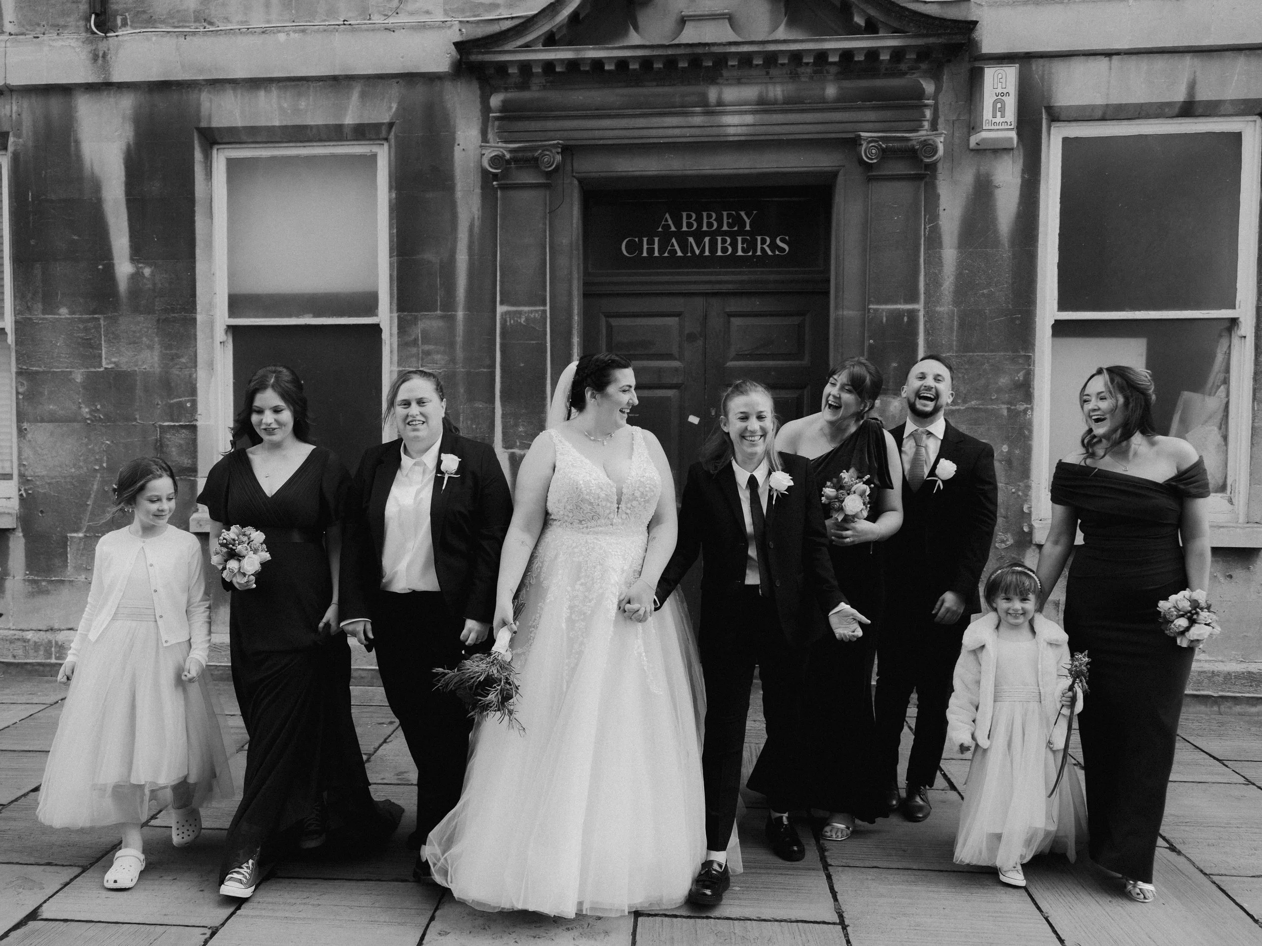 A black and white image of two brides, along with their bridal party, outside Abbey chambers in Bath city centre.