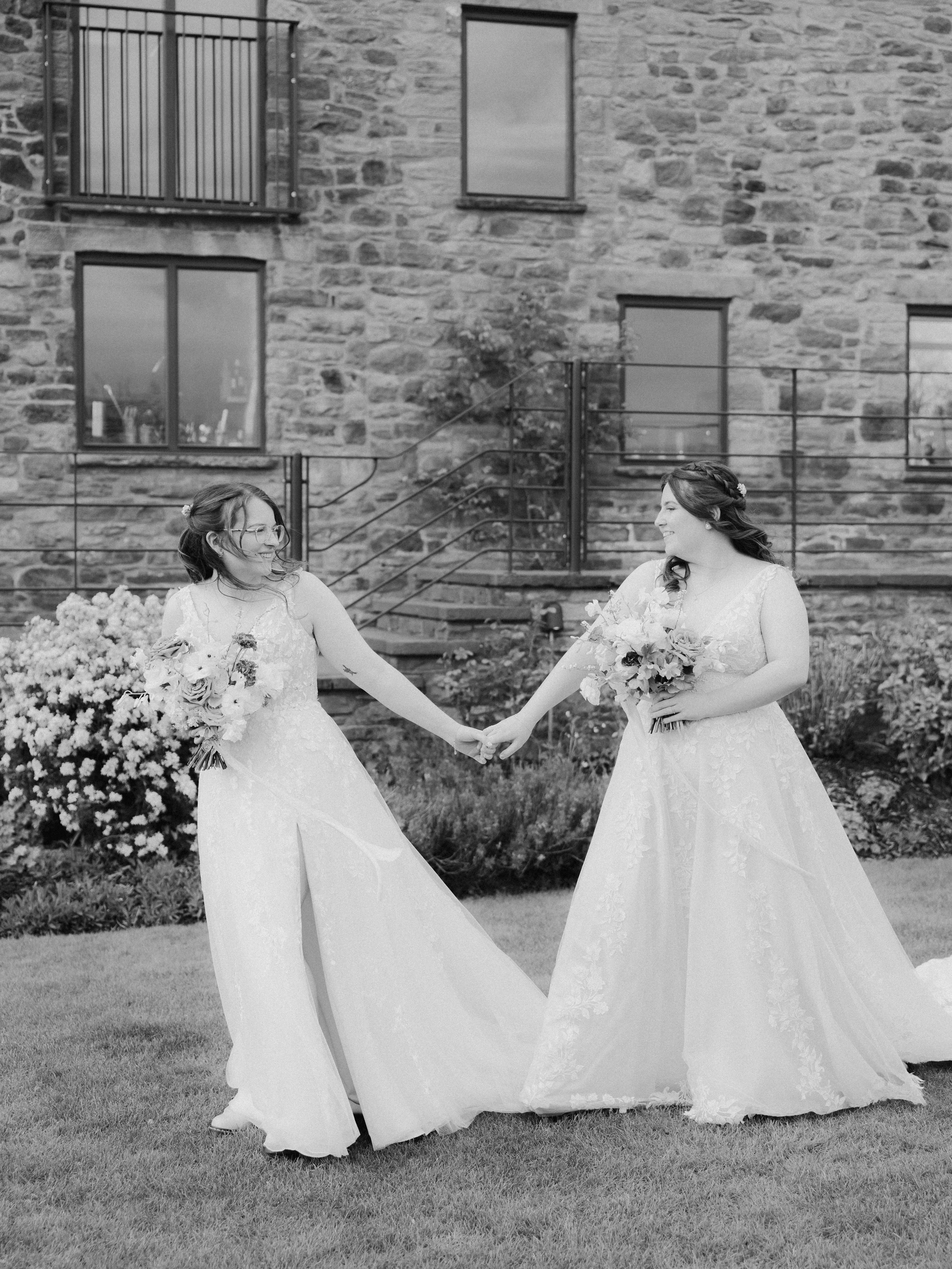 Two brides hold hands and smile at each other during couple portraits outside Llantilio House, with the stone house as a backdrop.