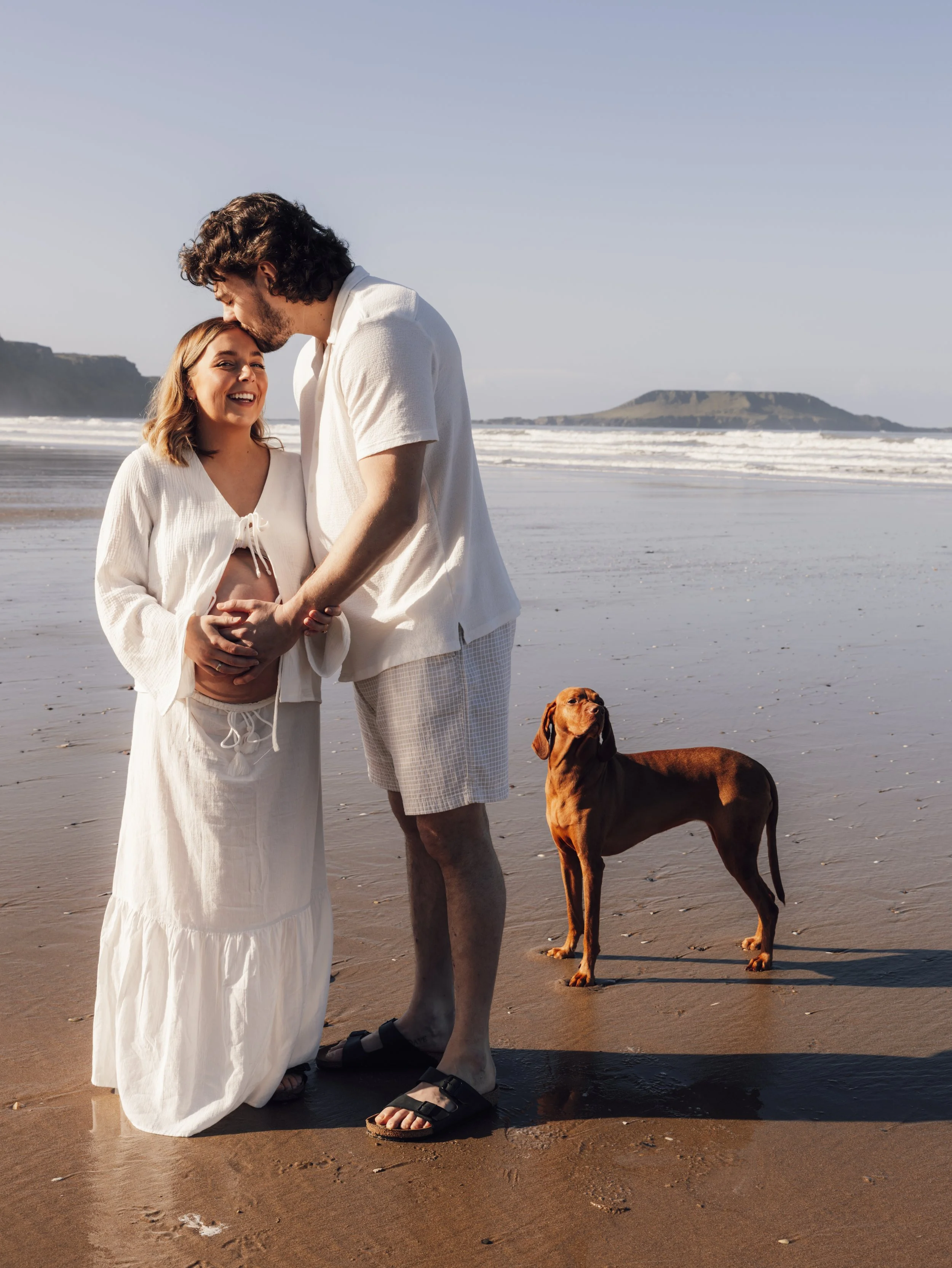 Pregnant couple walking along a South Wales beach with their dog at sunset