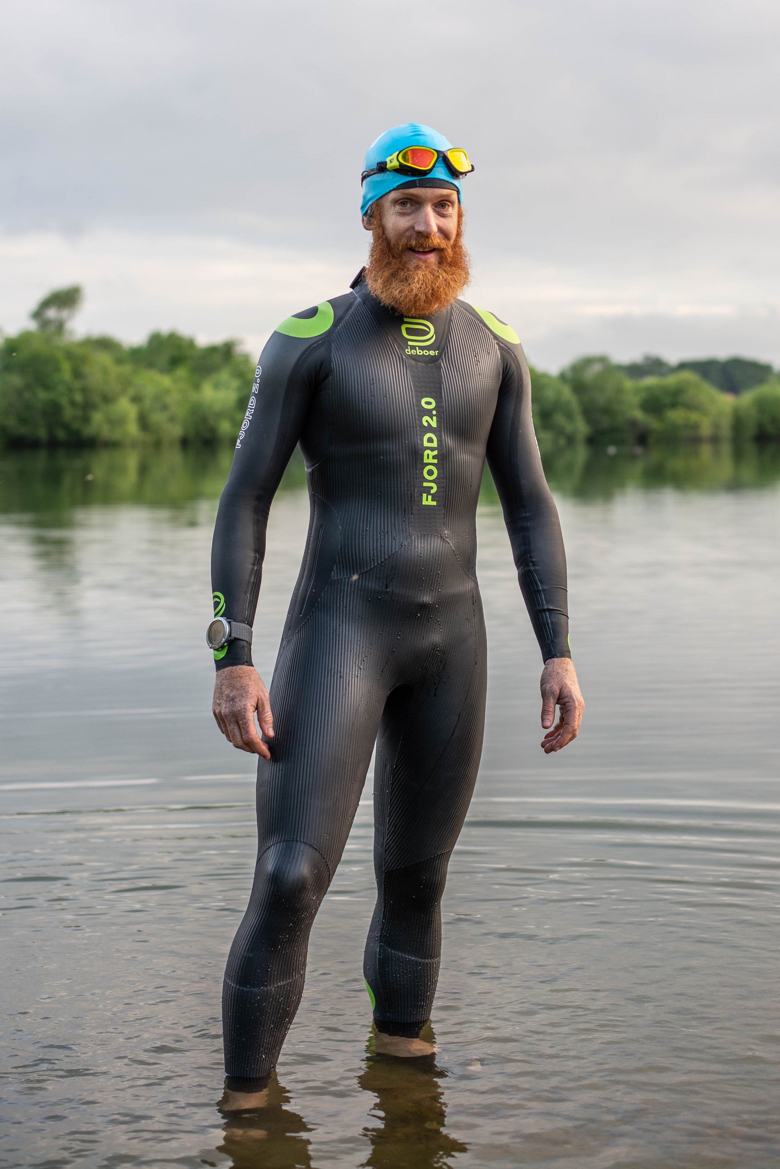 A person standing in shallow water wearing a black and green wetsuit, blue swim cap, and goggles. There are trees in the background.