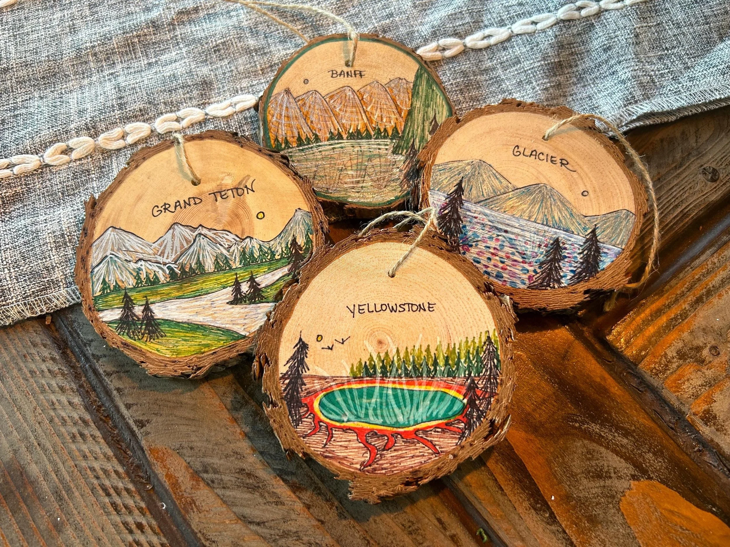 Four wooden slices with hand-painted mountain landscapes and labels of iconic Yellowstone National Park locations, including Grand Teton, Banff, Glacier, and Yellowstone with colorful trees and lakes painted on each slice.