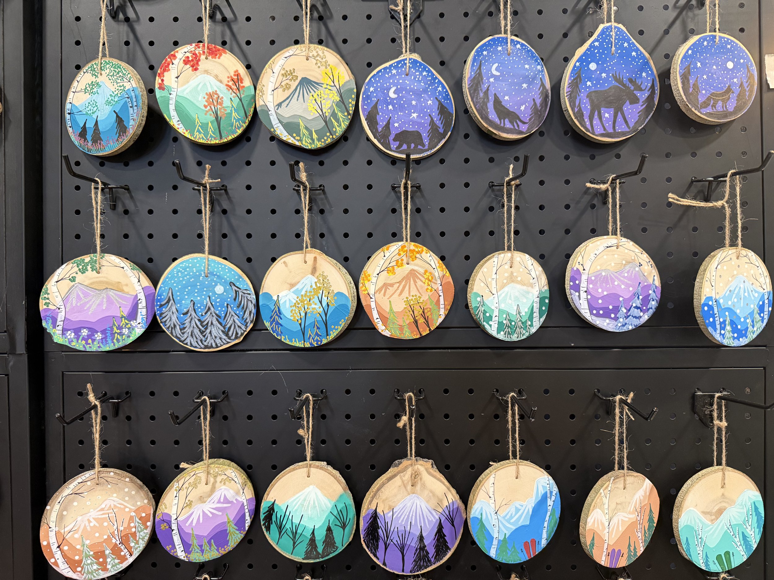 Set of painted wood slices depicting various landscapes including mountains, forests, animals, and night skies, hanging on a black display wall.